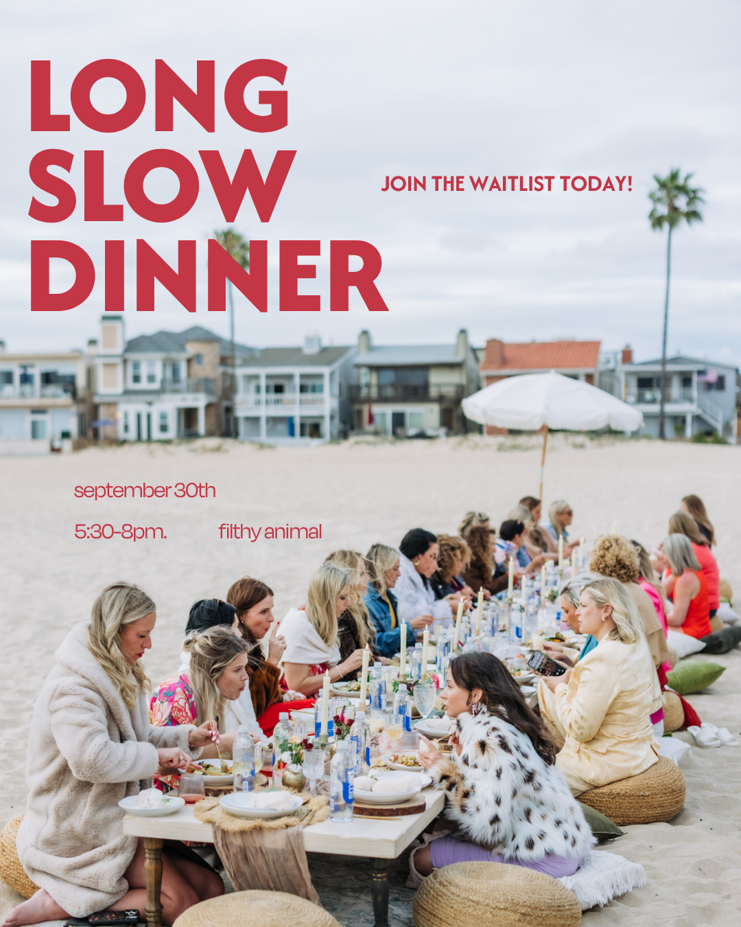 A group of people sitting on the beach having a meal at a long table with candles, surrounded by pillows and casual attire. Houses and palm trees are in the background under a cloudy sky.