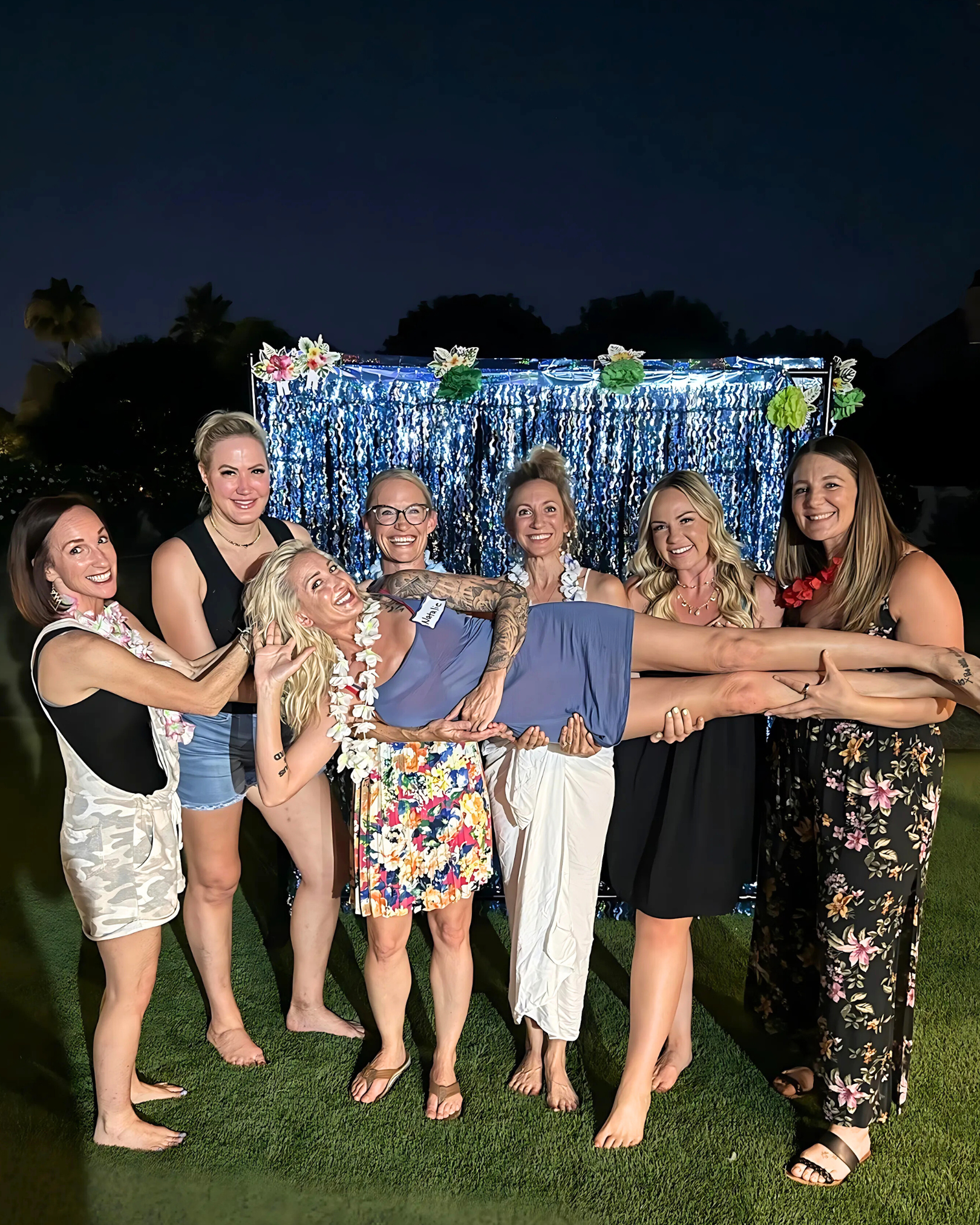 Group of women at a nighttime outdoor party, with one woman being held horizontally by the others, in front of a blue Tiki backdrop decorated with flowers and greenery.