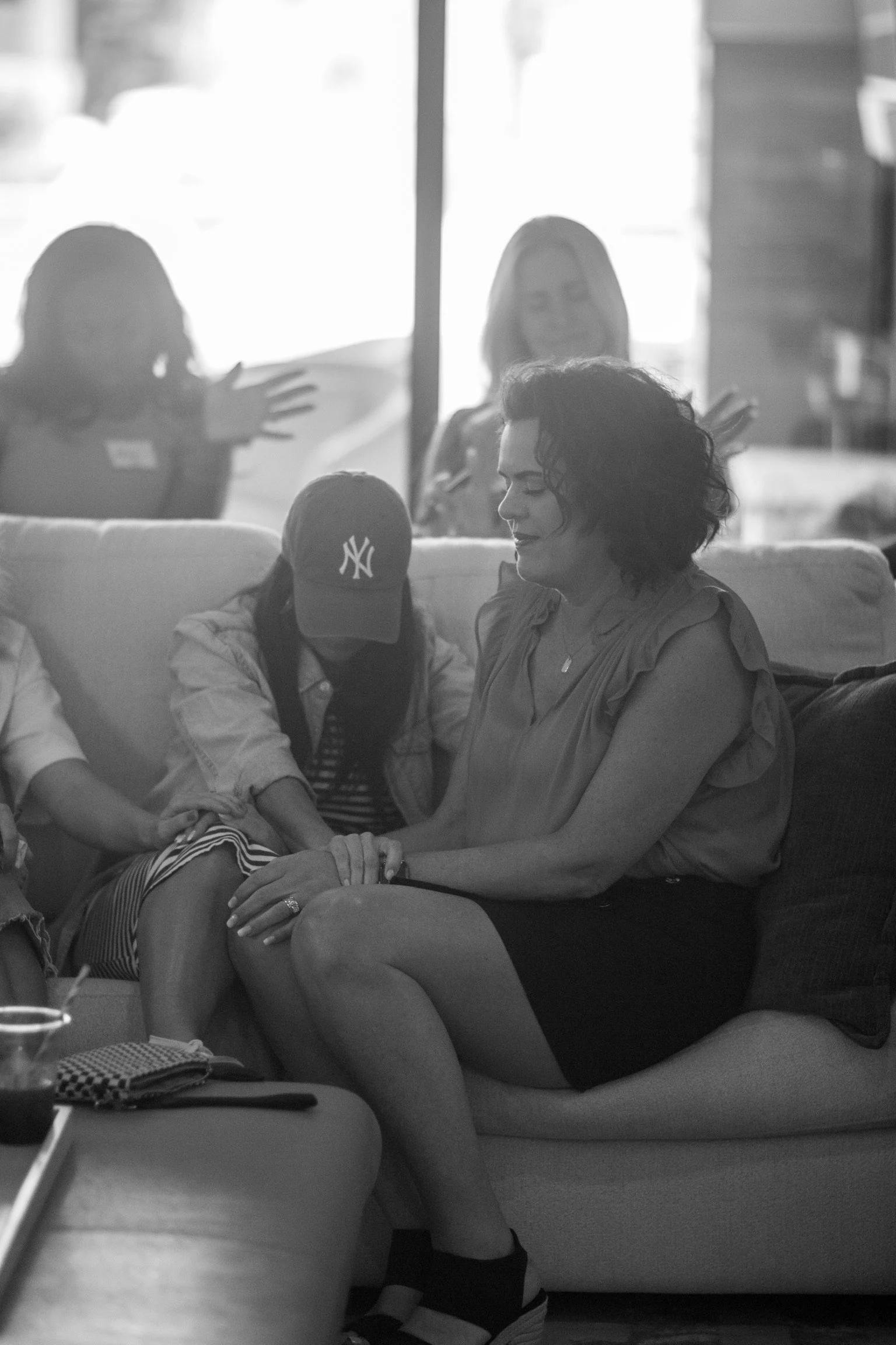 A woman with curly hair sitting on a couch, holding a young girl in a white jacket with a New York Yankees cap, while outside through the window, other women are talking or laughing.