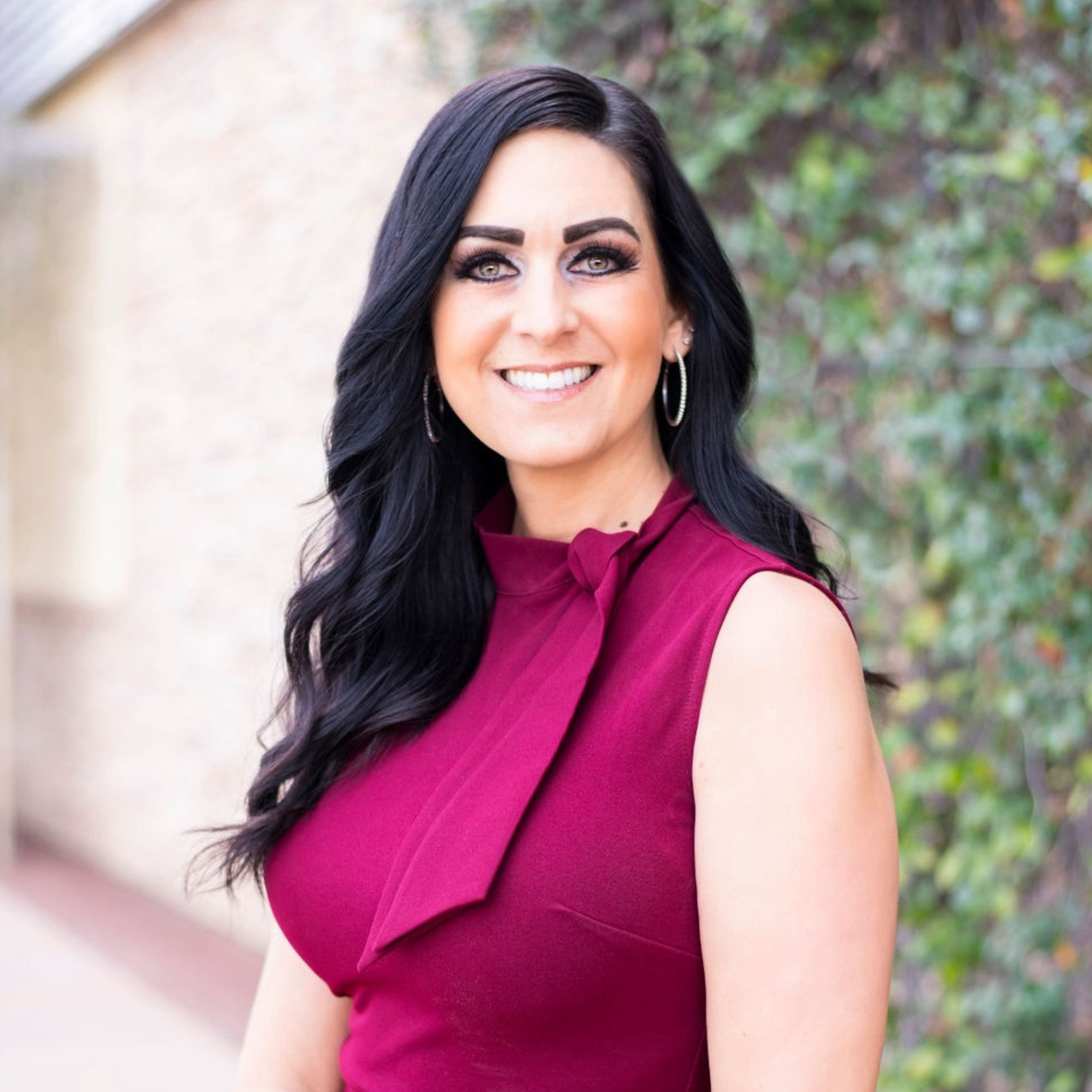 A woman with long black hair, wearing a sleeveless red dress with a bow detail, standing outdoors with greenery in the background and smiling.
