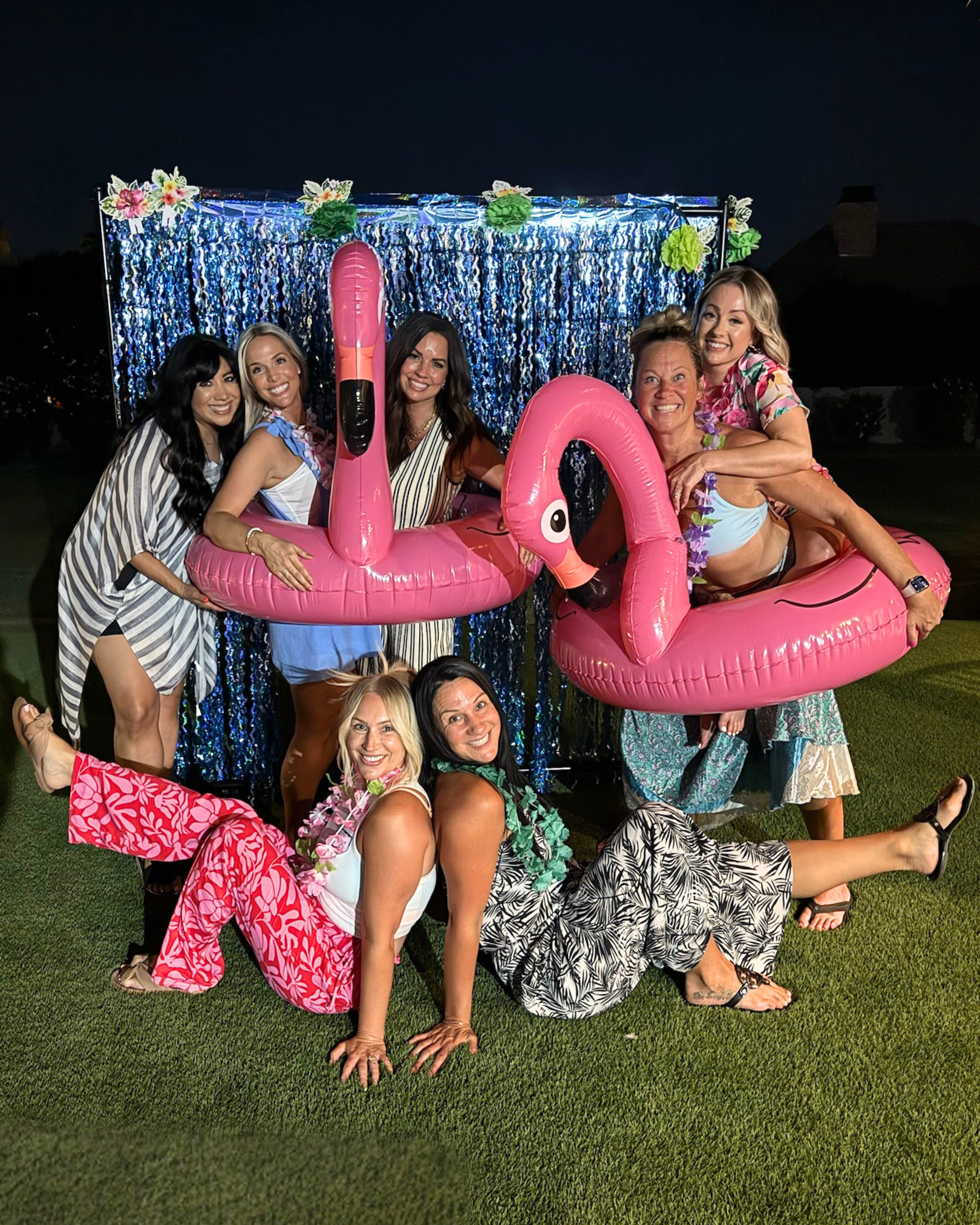 Group of seven women at a party, some wearing leis, posing with inflatable pink flamingos in front of a shiny blue and silver backdrop at night.