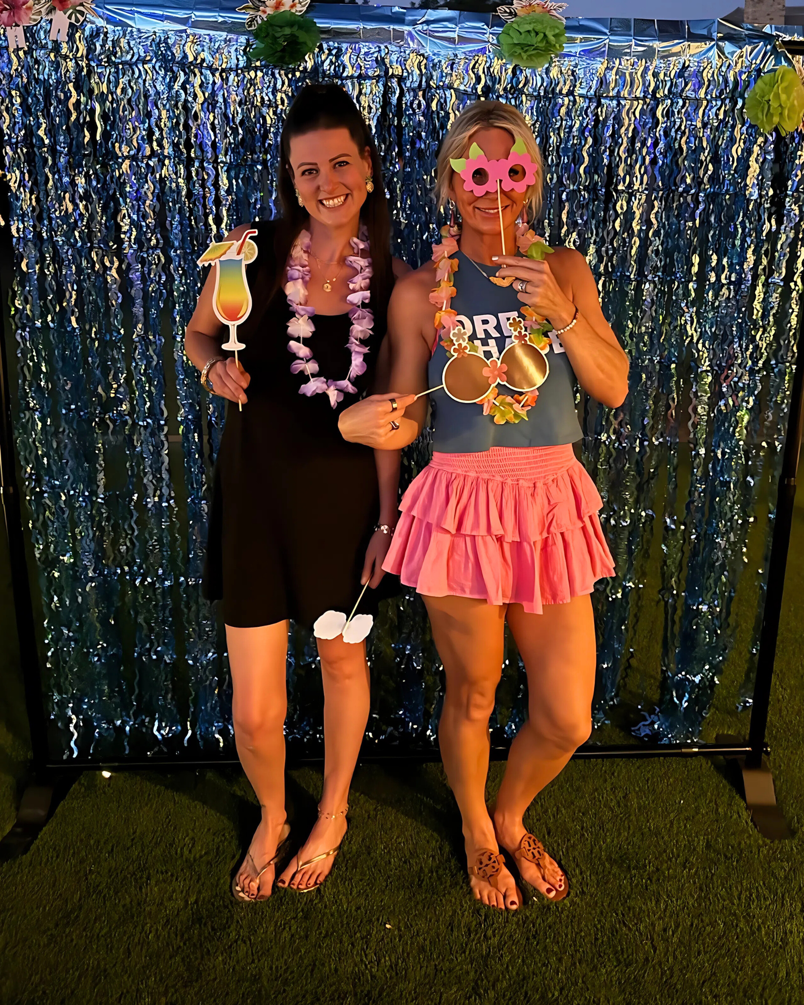 Two women standing in front of a shiny blue and silver backdrop, holding fun party props, wearing leis, and dressed in colorful summer attire, suggesting a festive or tropical-themed party.