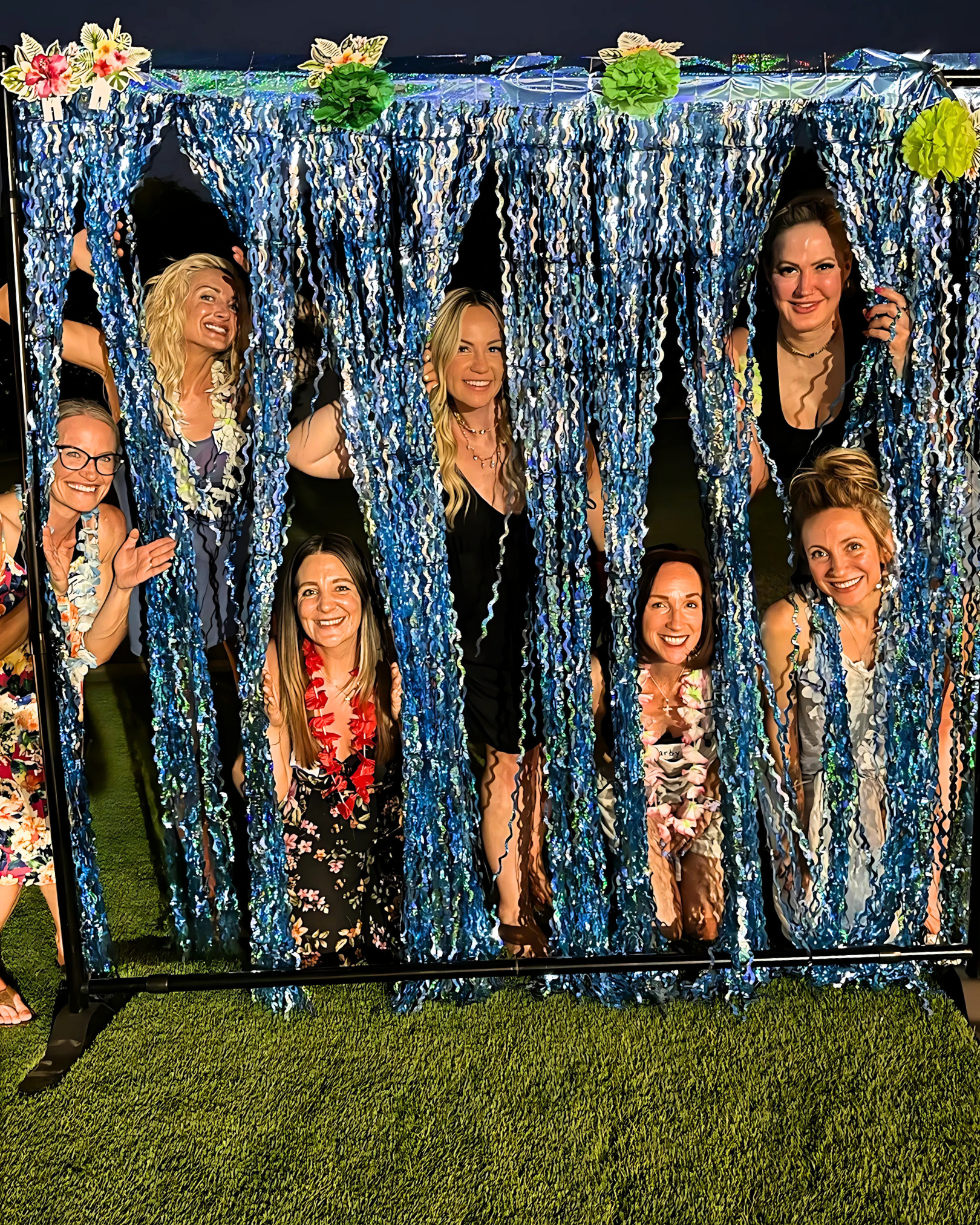 Group of women at a party posing behind a blue metallic fringe curtain decorated with flower leis and paper flowers, standing and smiling on green grass.