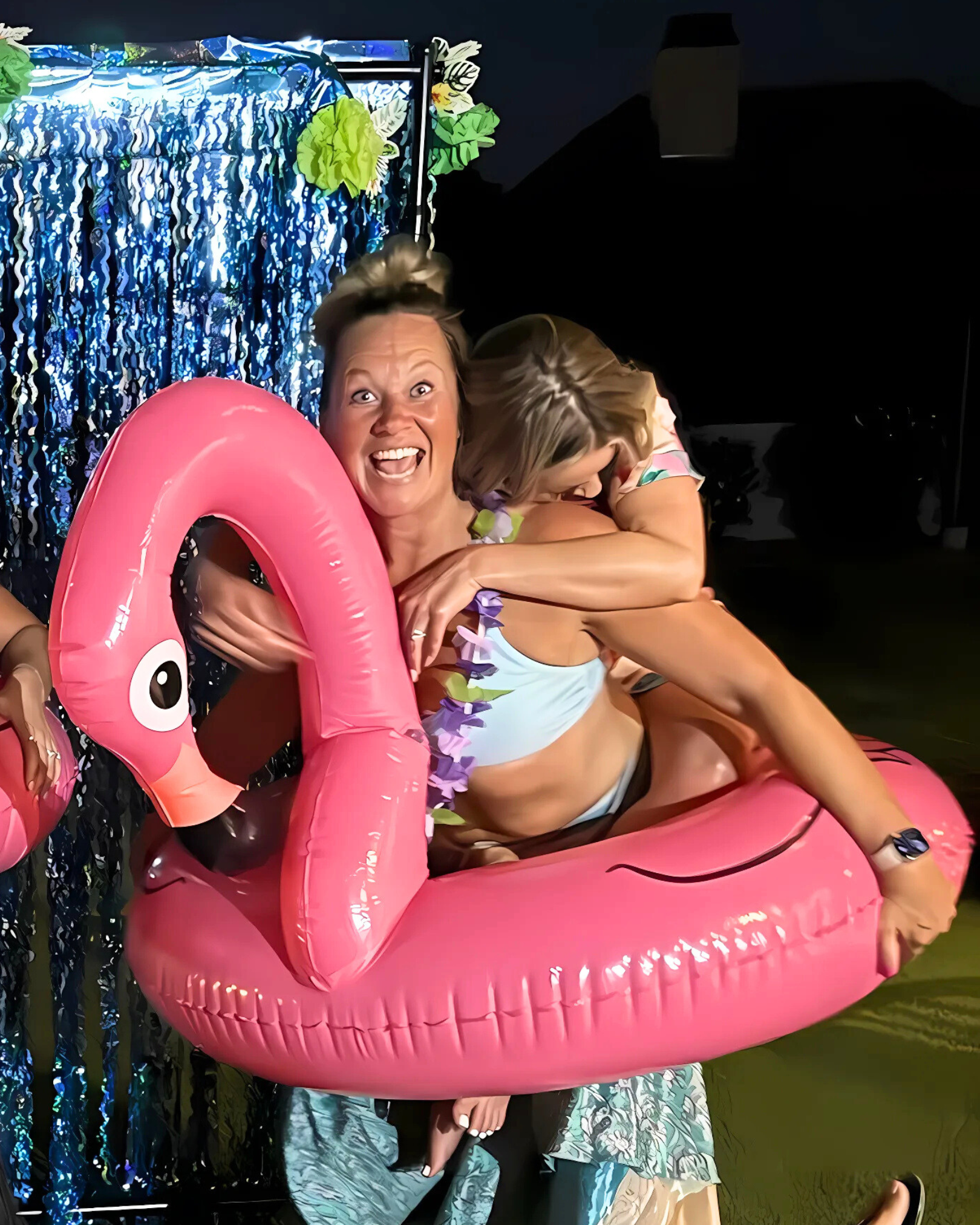Two women on a pink flamingo pool float, one smiling excitedly while the other is hugging her from behind, in a festive outdoor setting at night with a colorful backdrop.