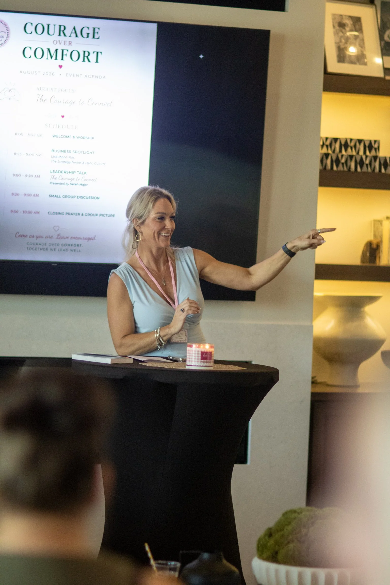 A woman standing inside near a large screen displaying an event schedule titled 'Courage Over Comfort.' She has blonde hair, wears a light-colored sleeveless dress, large earrings, and a wristwatch, and is smiling while gesturing with her hand. In th