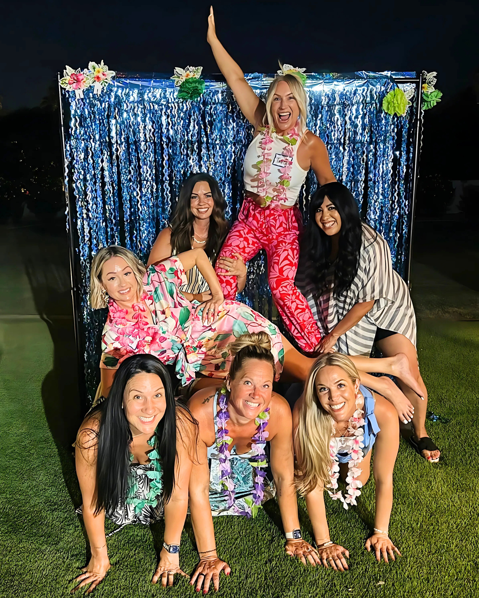 Group of women participating in a limbo dance outdoors at night, with a blue fringe backdrop and tropical flowers, wearing floral shirts and leis, smiling and having fun.