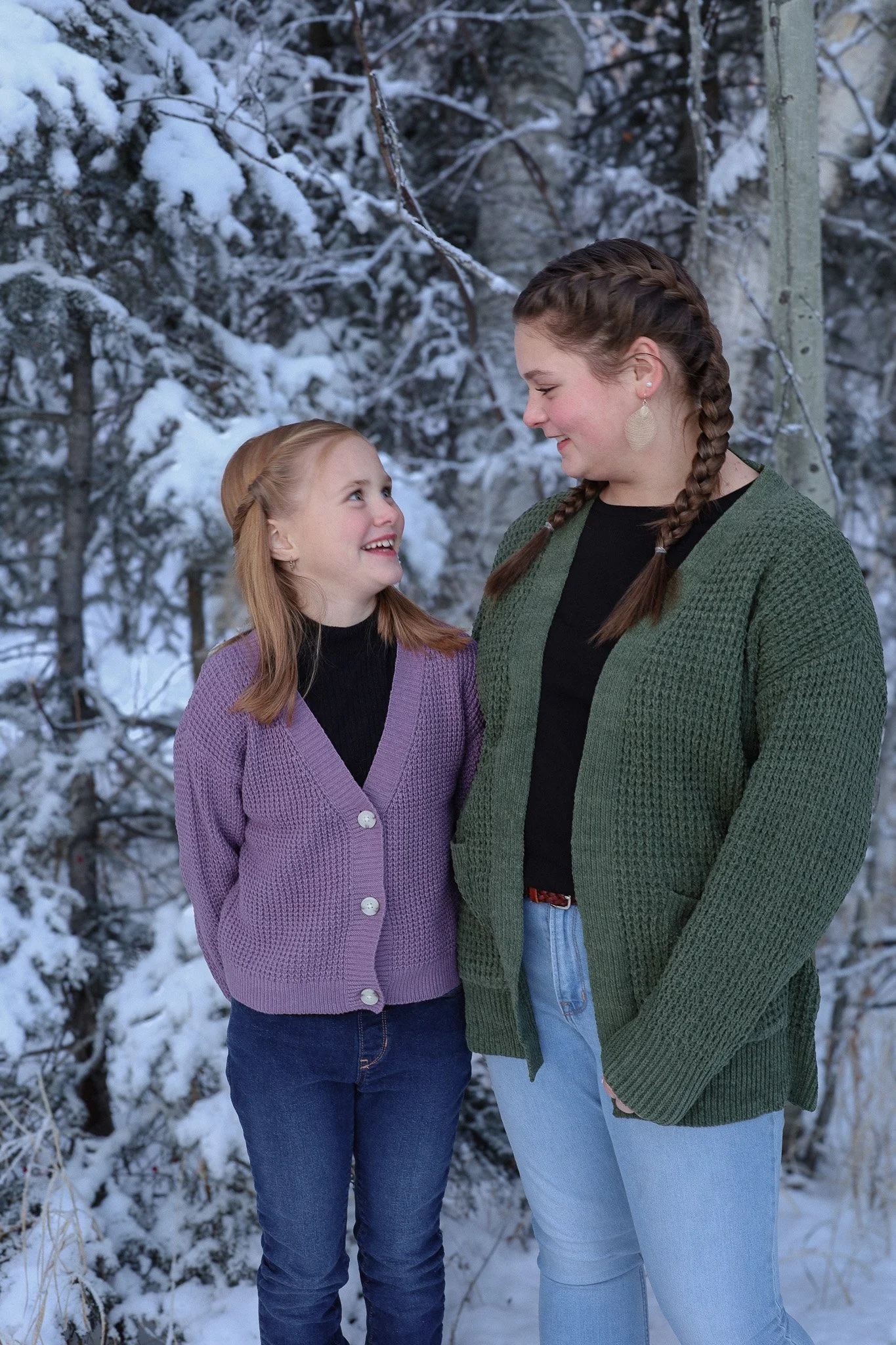 A young girl and an older girl or woman standing in a snowy wooded area, smiling and looking at each other.