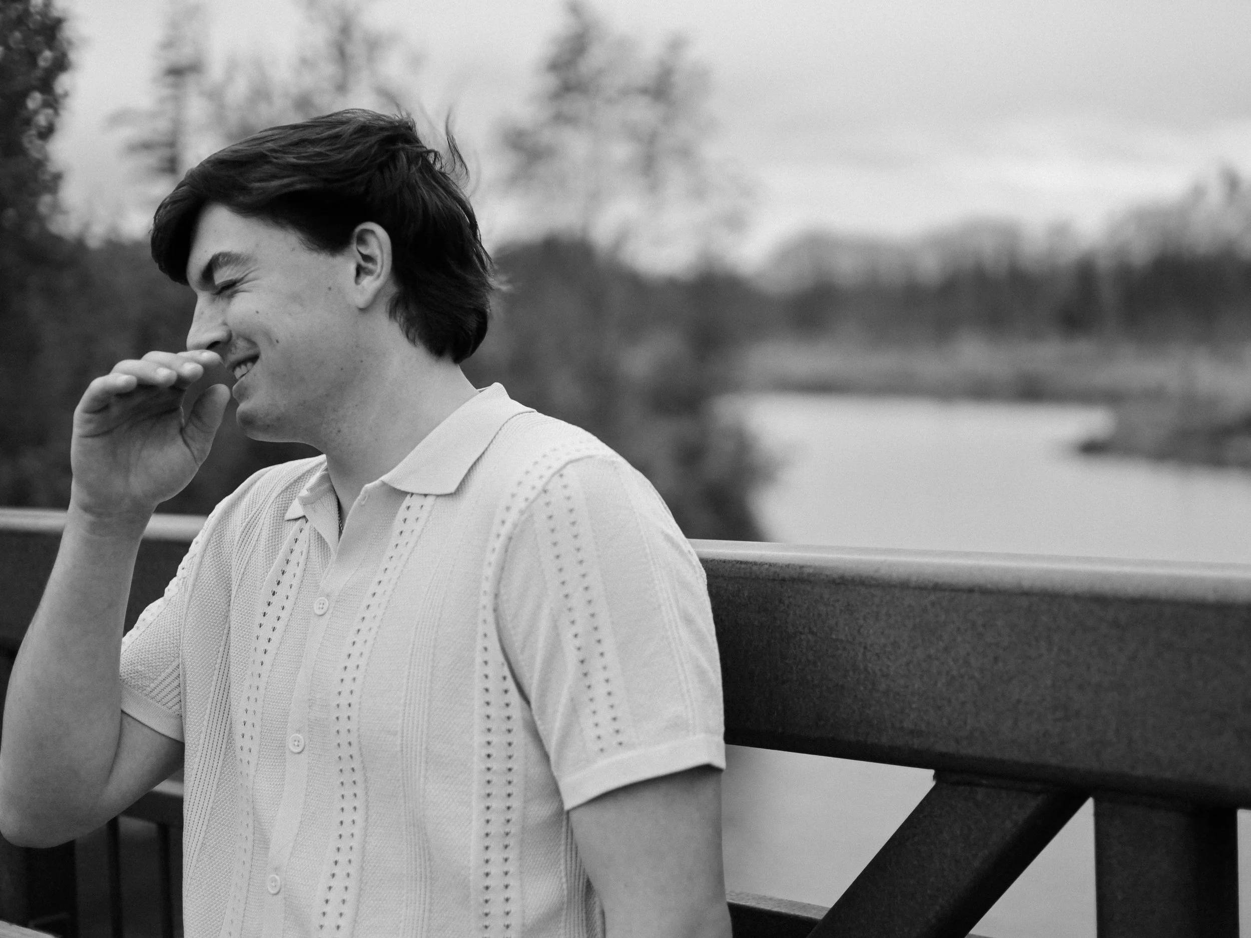 A young man with dark hair smiling and laughing outdoors near a river, wearing a light-colored short-sleeve shirt, captured in black and white.