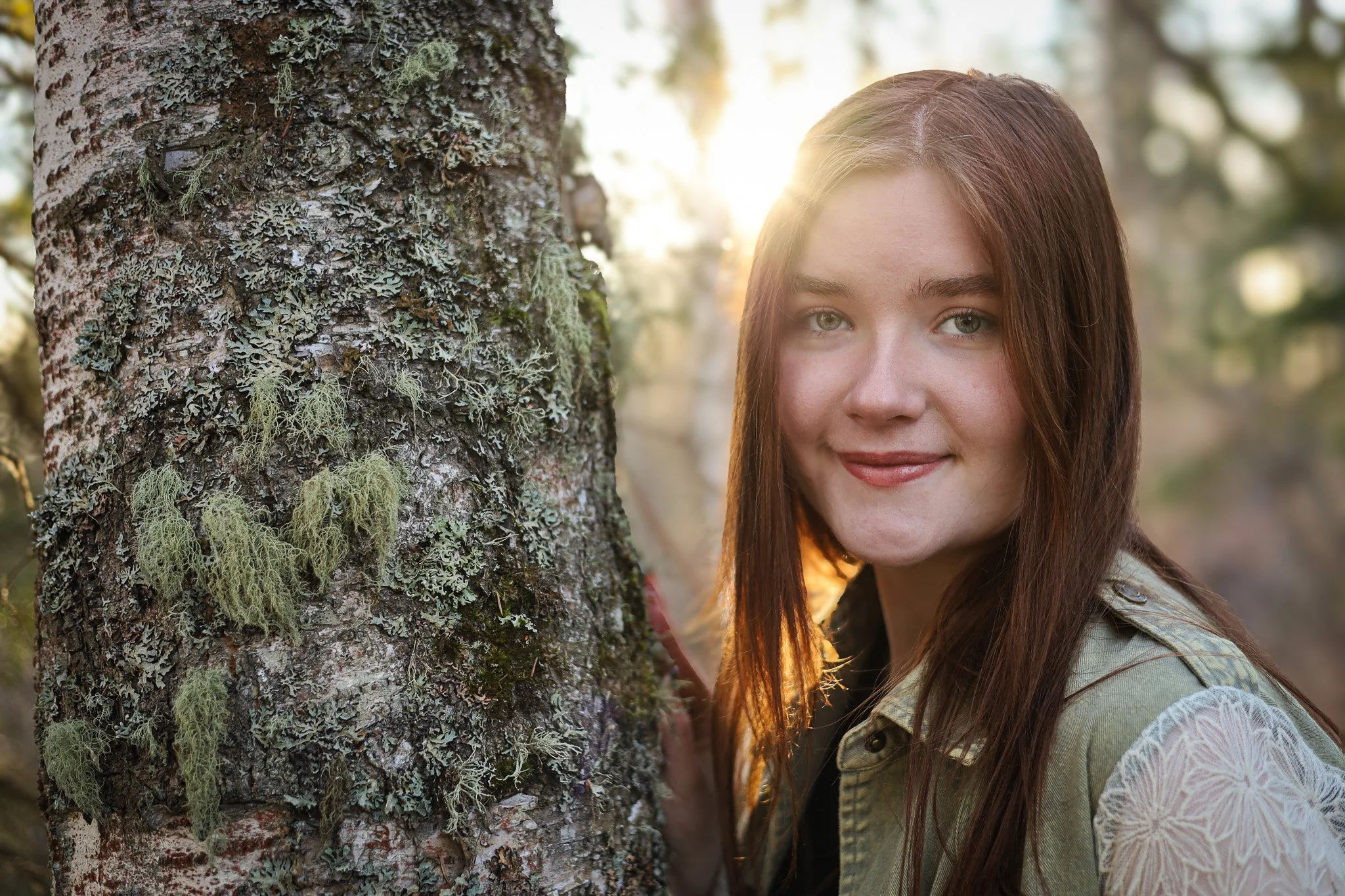 Young woman with long reddish-brown hair smiling outdoors near a tree with moss and lichen, backlit by the sun.