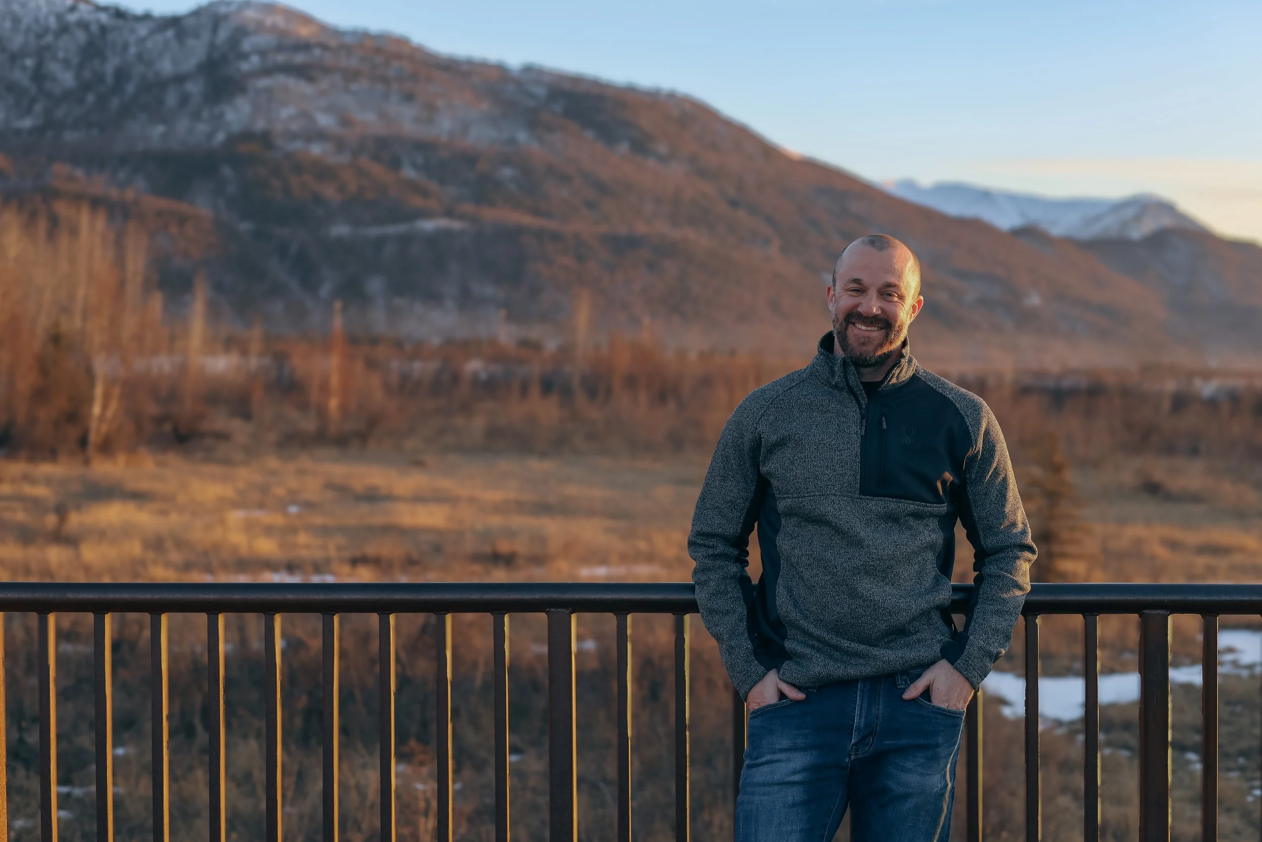 A smiling man with a beard and short hair standing outdoors in front of a mountain landscape during sunset, wearing a gray and black jacket with his hands in his pockets.