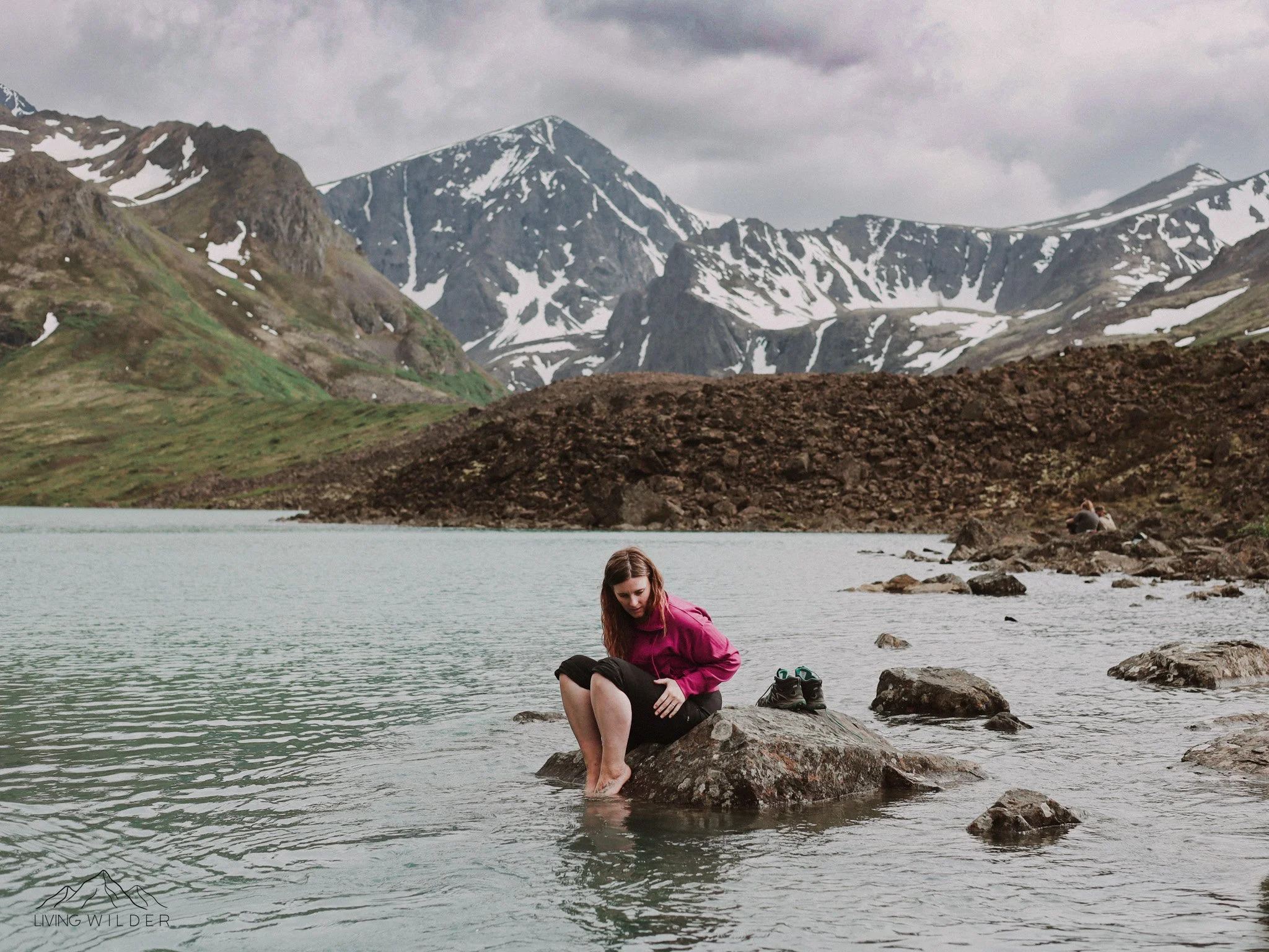 Woman sitting on a rock in a lake surrounded by mountains with snow, some green slopes, and cloudy sky.