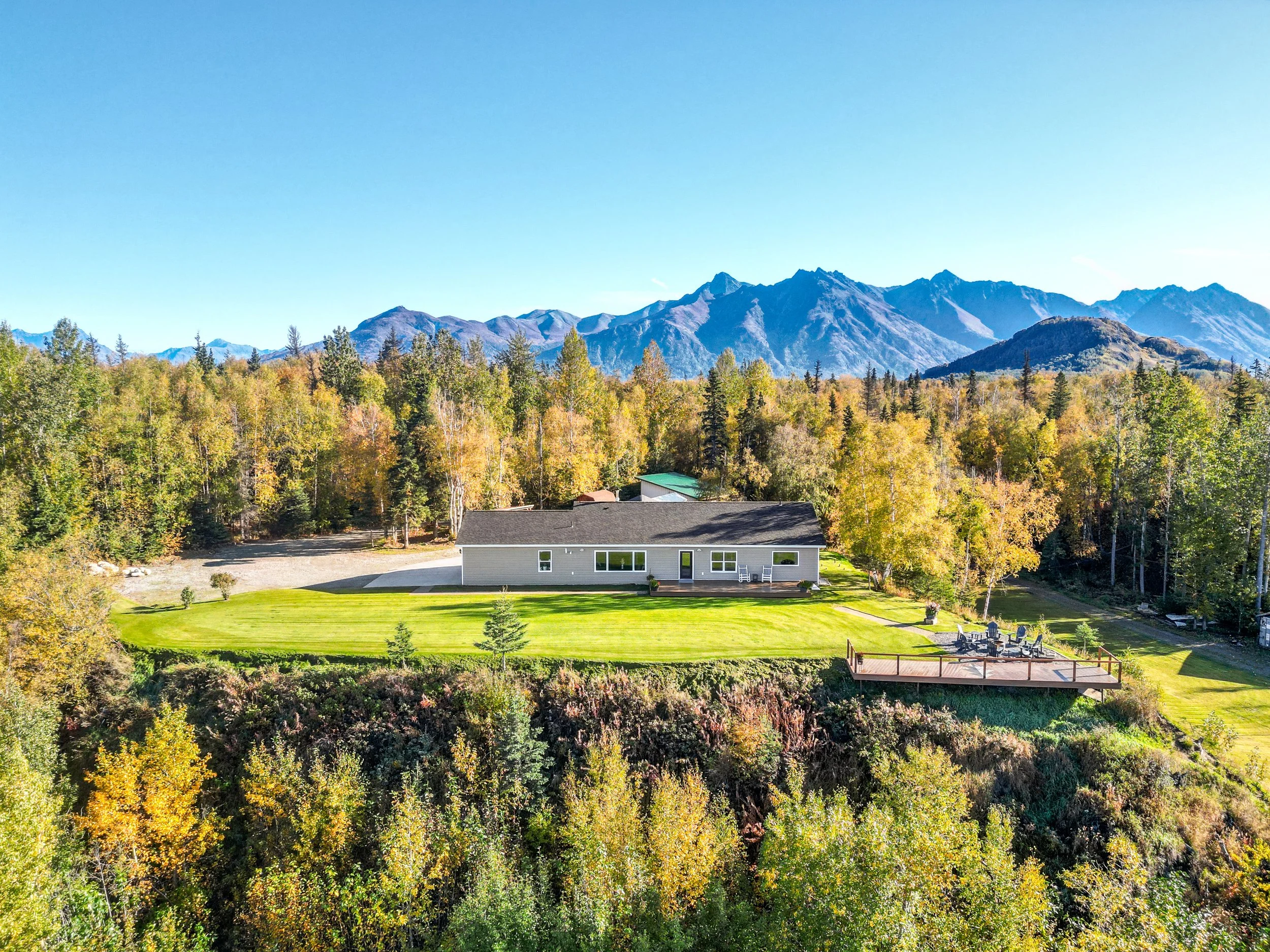 A house with a well-maintained lawn and a deck, surrounded by trees with fall foliage, and mountains in the background under a clear blue sky.