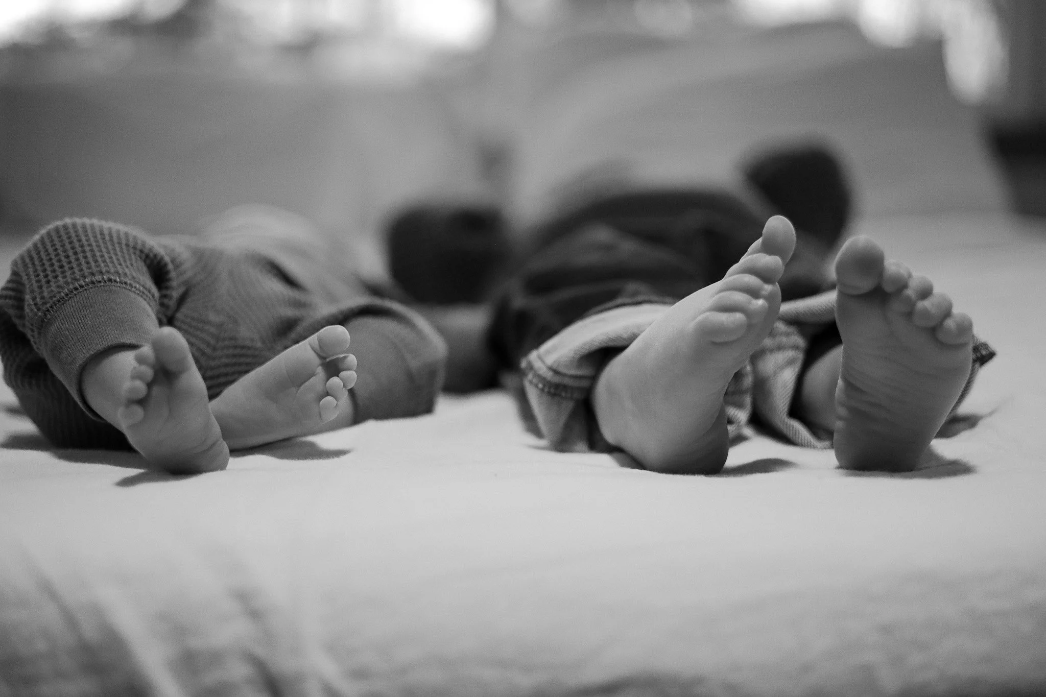 Black and white photo of two baby dolls lying on their backs on a soft surface, showing close-up of their feet, with their legs slightly bent.