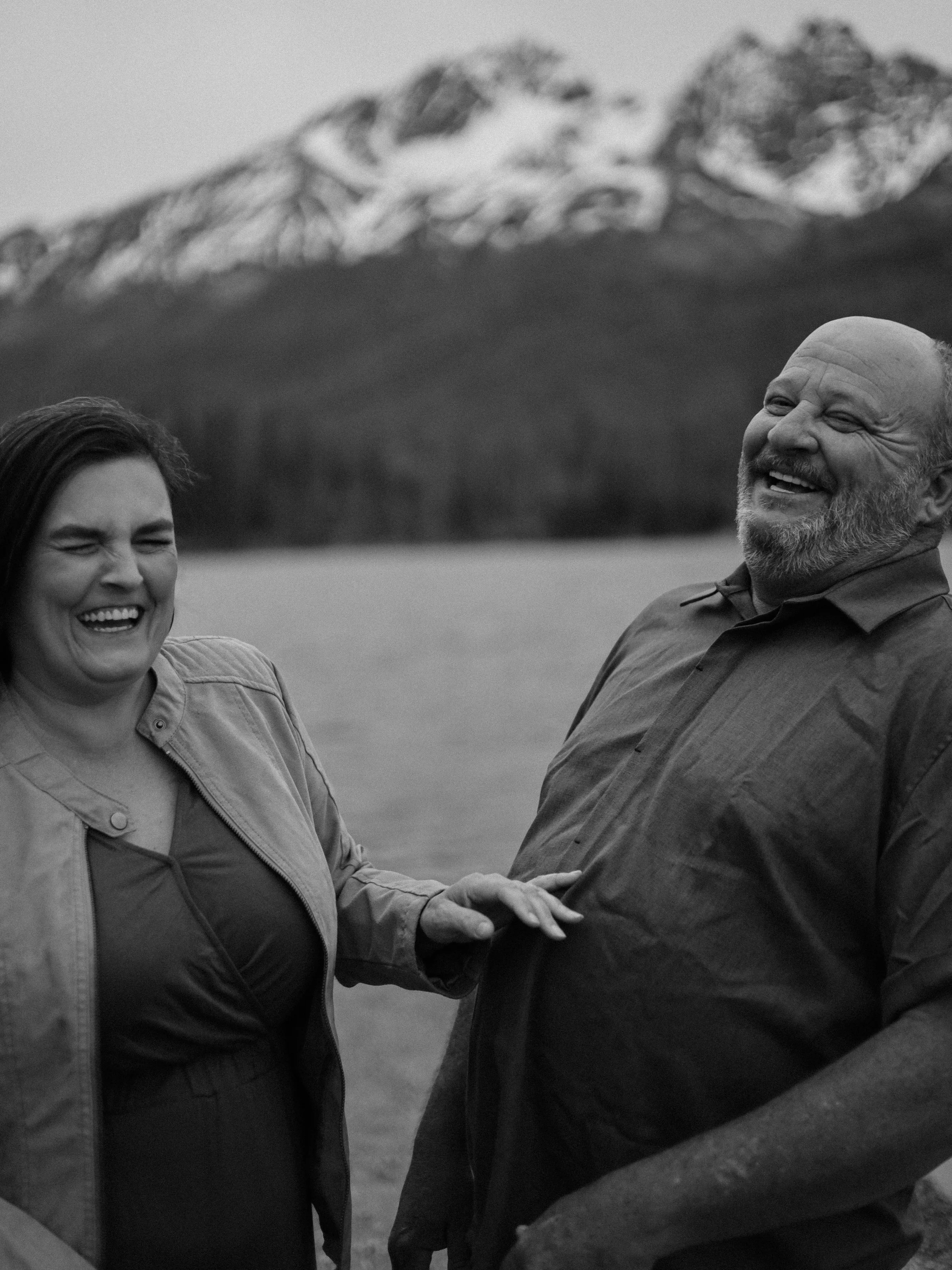 A black-and-white photo of a woman and man laughing outdoors near a lake with snow-capped mountains in the background.