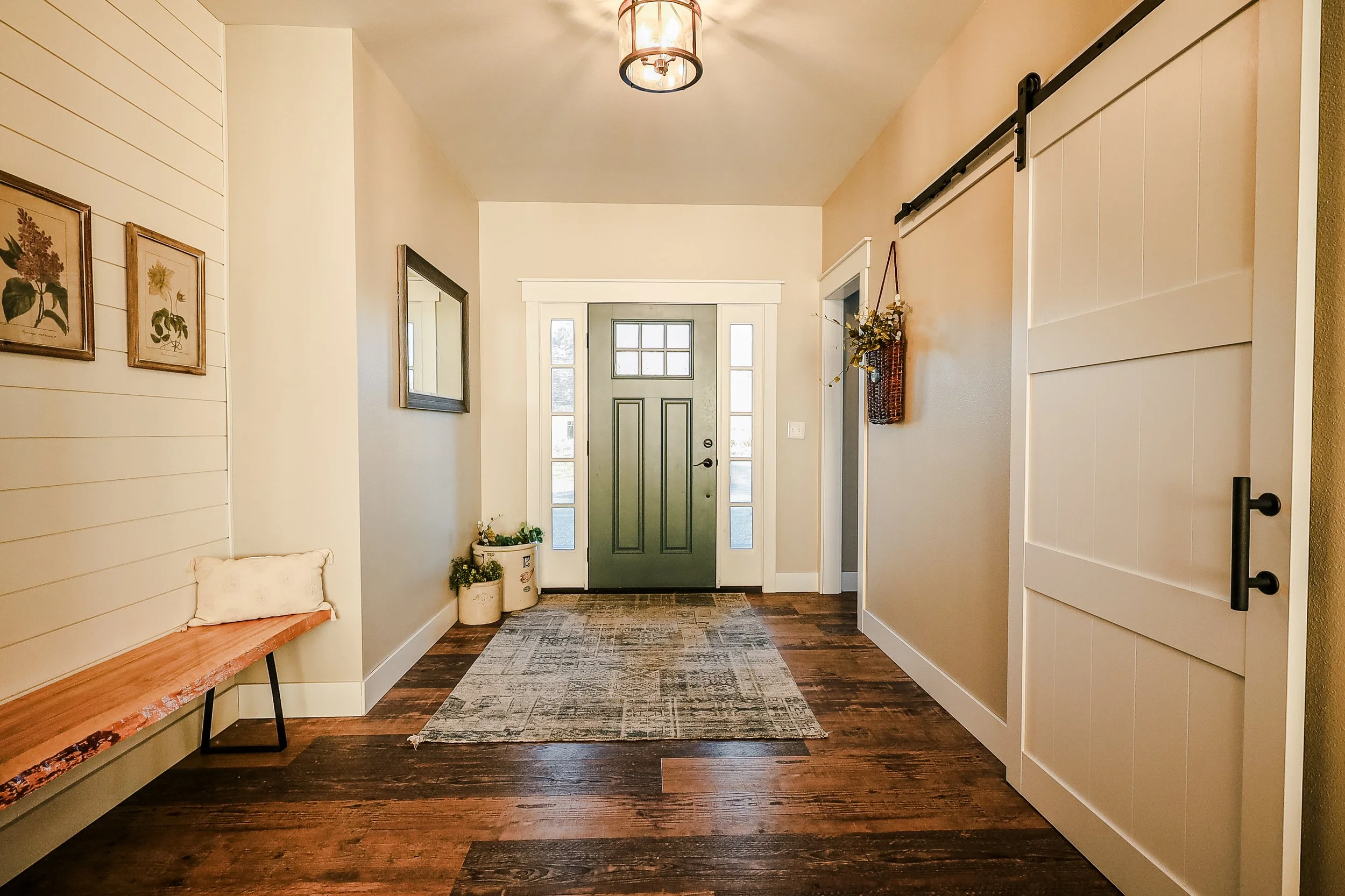 Entryway with hardwood floors, a green front door with windows, a bench on the left, and decorative plants and wall art.