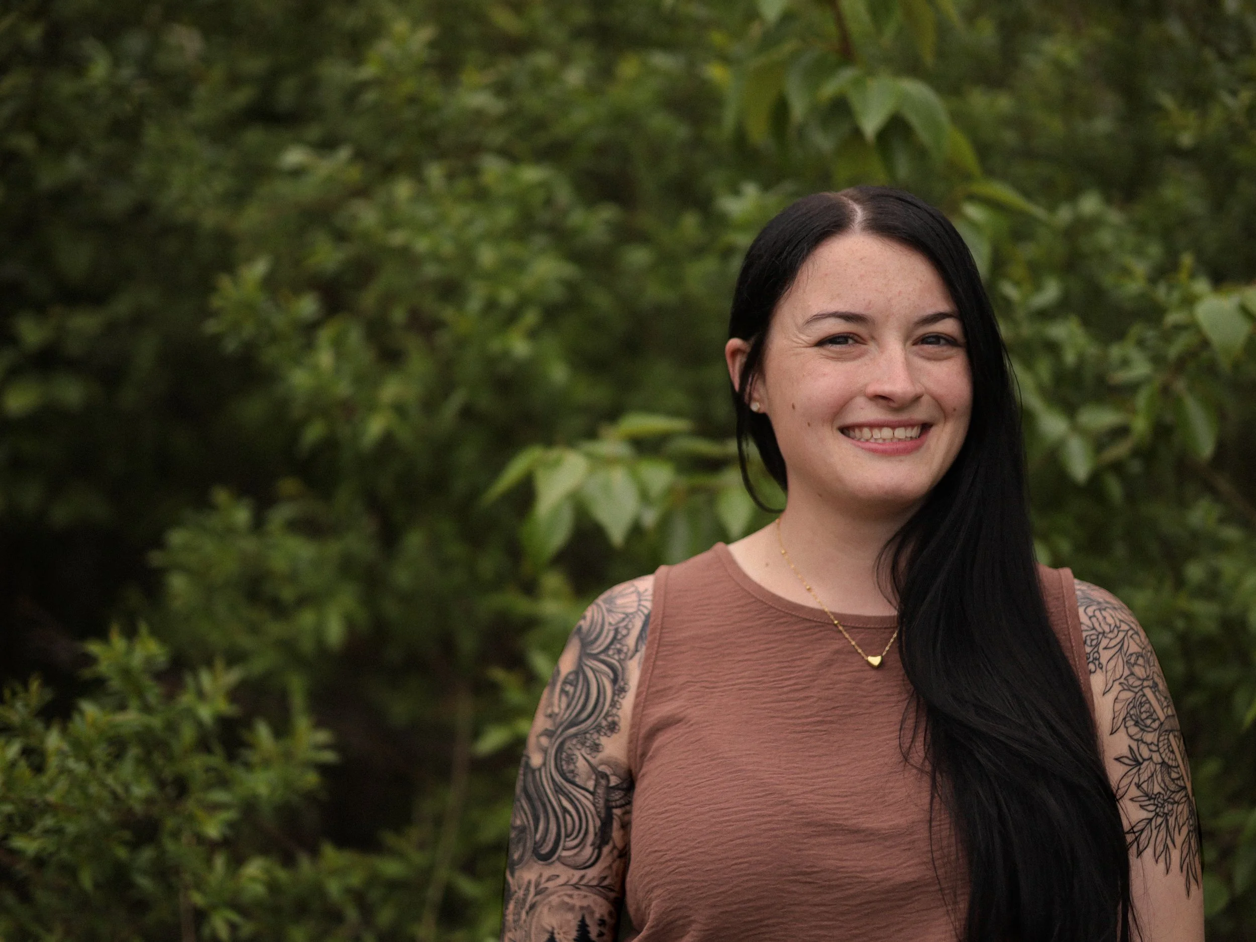 A woman with long black hair, wearing a sleeveless brown top and a gold necklace, smiling outdoors with green foliage in the background.