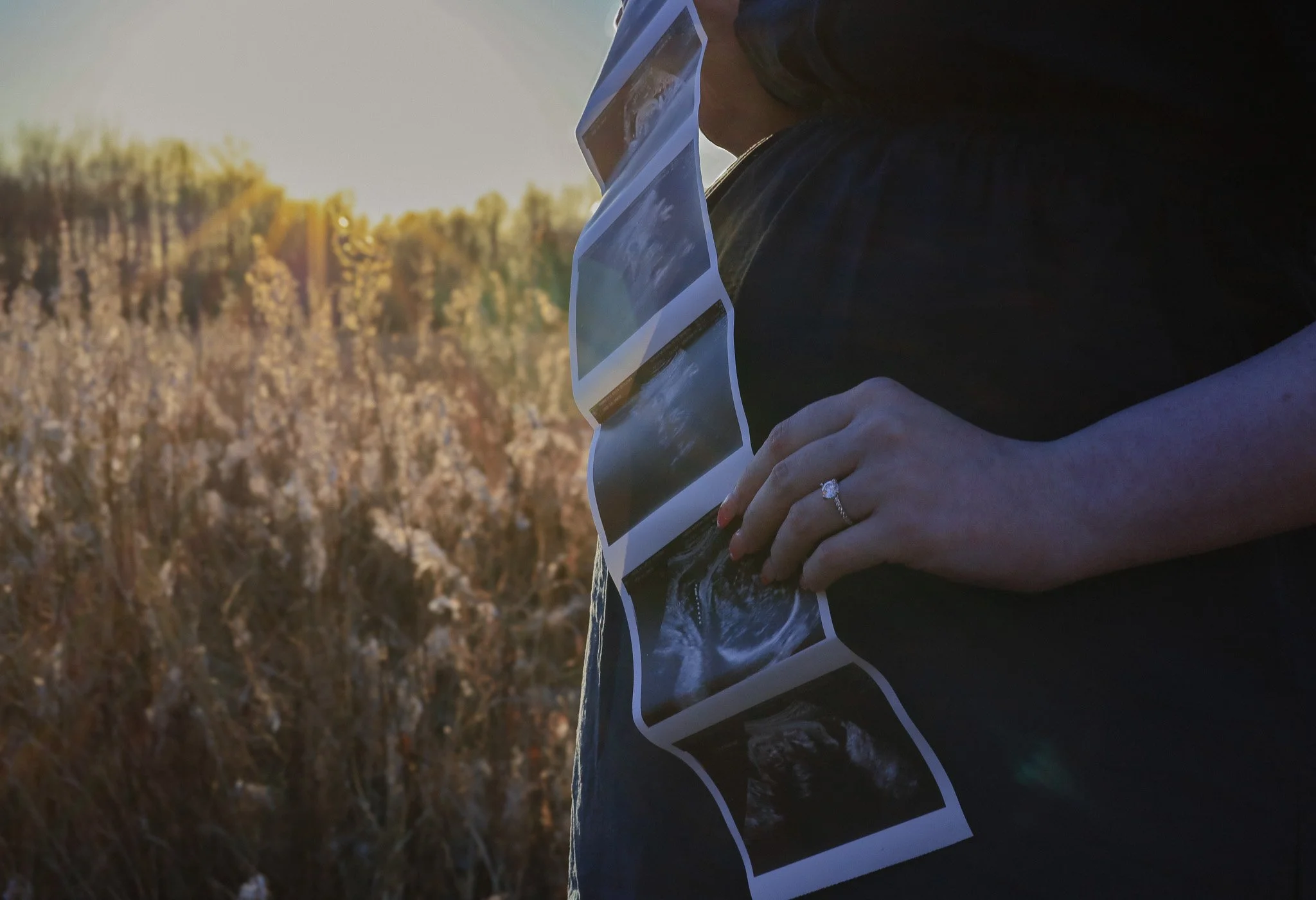 Person holding ultrasound photo prints outdoors at sunset or sunrise, with a field and treeline in the background.