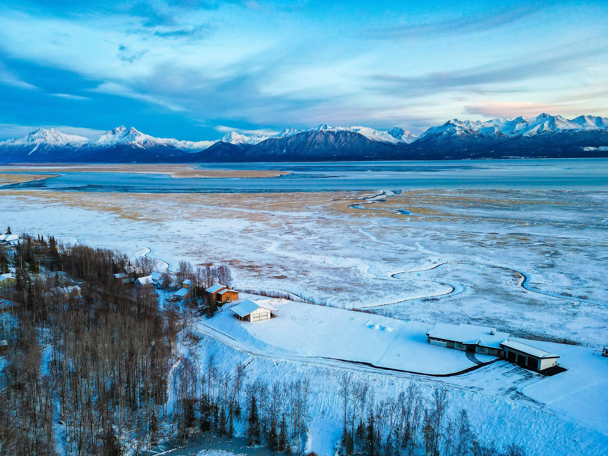 Snow-covered landscape with a few houses and trees in the foreground, a frozen body of water, and snow-capped mountains in the background under a partly cloudy sky.