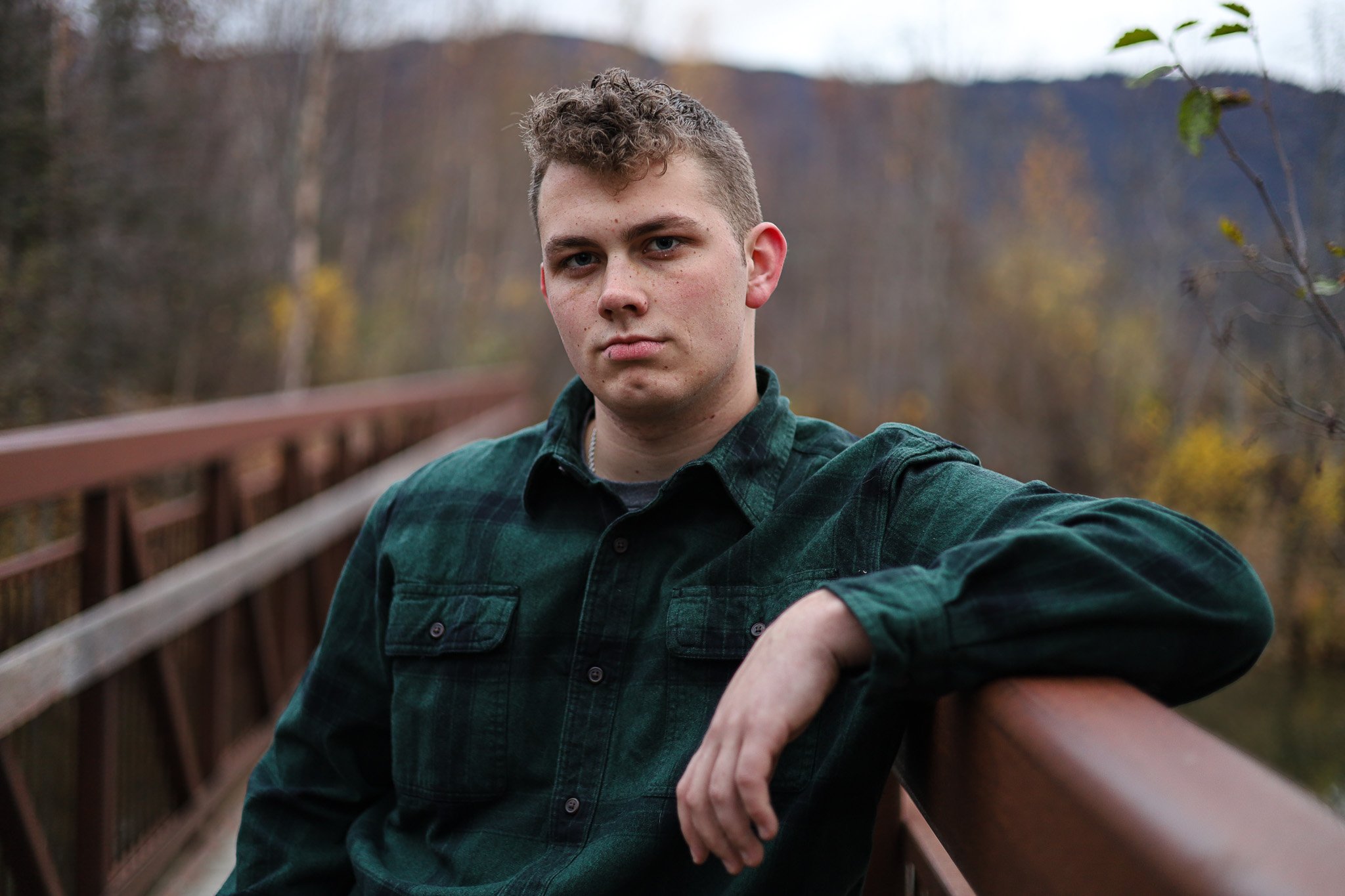 A young man with short curly hair standing on a wooden bridge, wearing a dark green flannel shirt, outdoors during autumn.