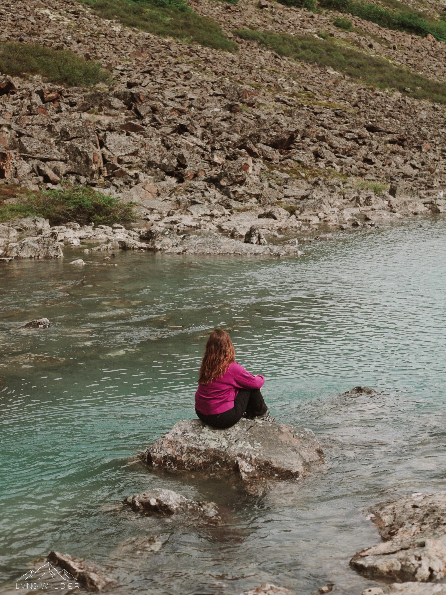 A woman with long hair sitting on a large rock at the edge of a calm mountain lake, surrounded by rocky terrain and sparse greenery.