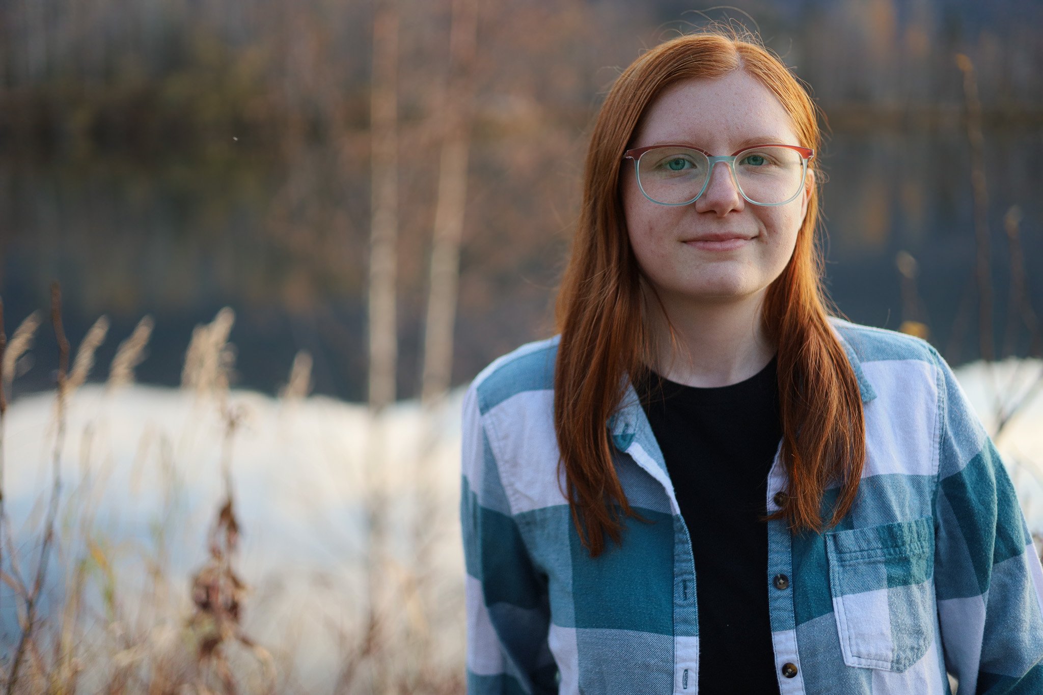 A young woman with red hair and glasses standing outdoors in a natural setting with trees and snow, facing the camera with a slight smile.