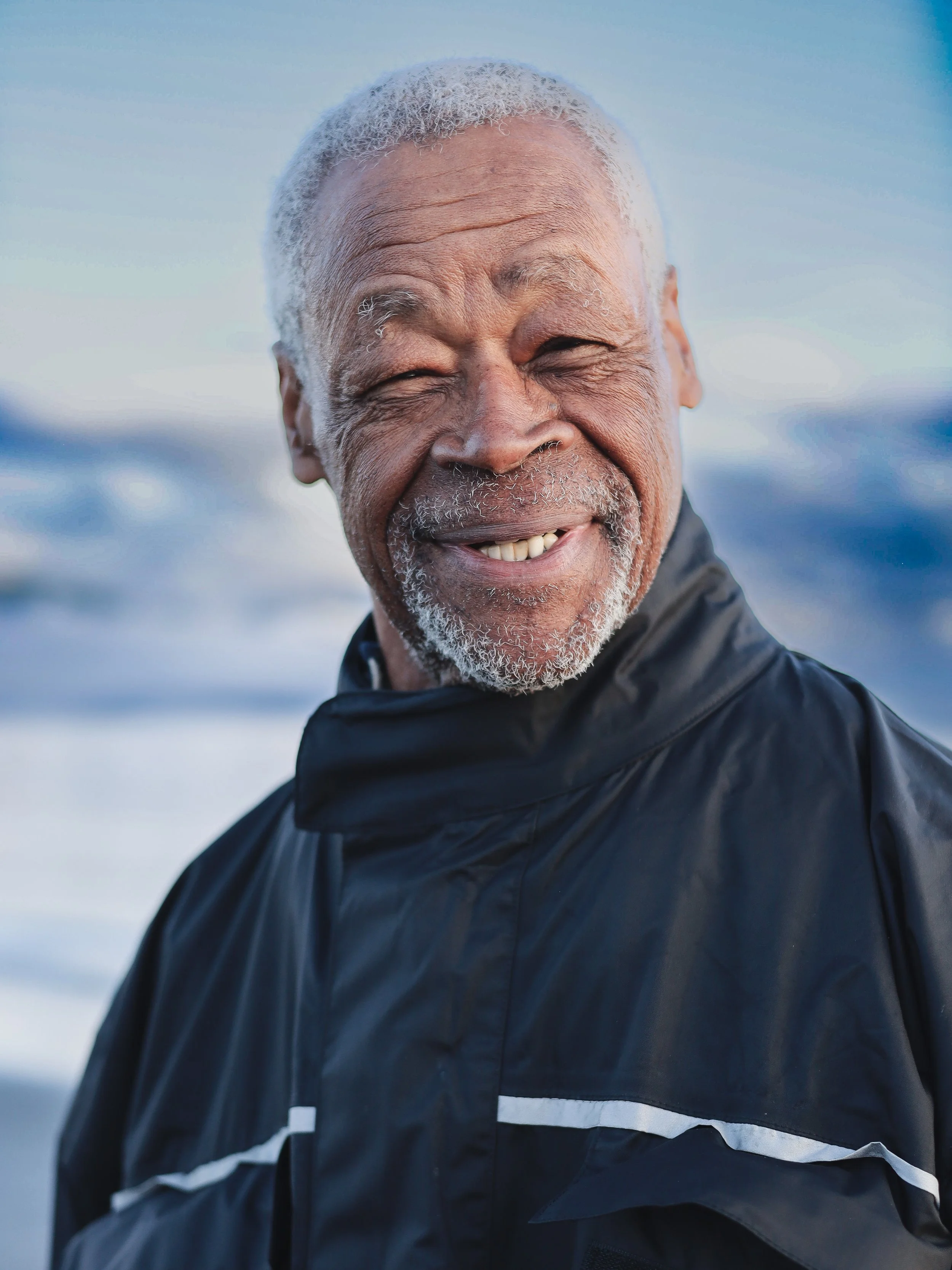 Close-up of an elderly man with gray hair and beard smiling outdoors, wearing a black waterproof jacket with a snowy background.
