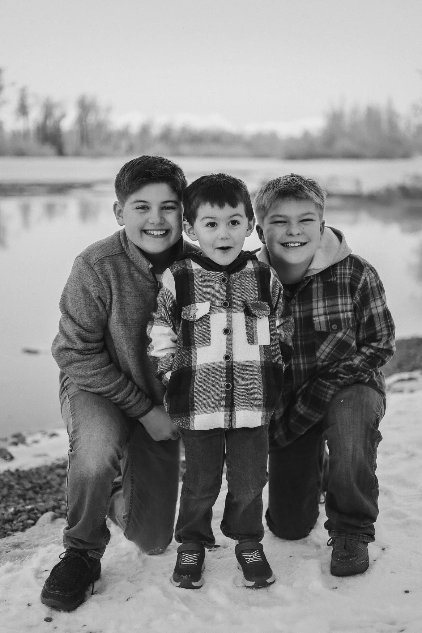 Three smiling boys, two crouching and one standing, outdoors in a snowy landscape near a body of water, black and white photograph.