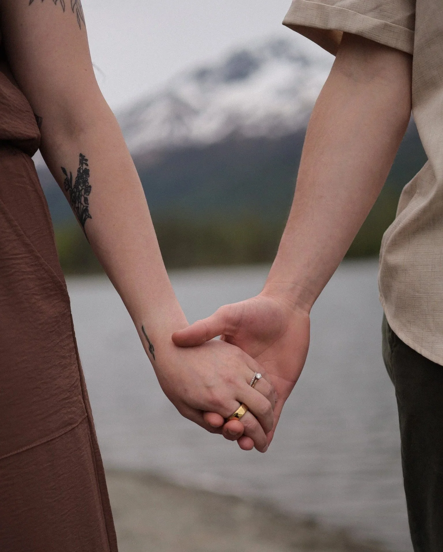 Close-up of a couple holding hands near a lake, with mountains in the background.