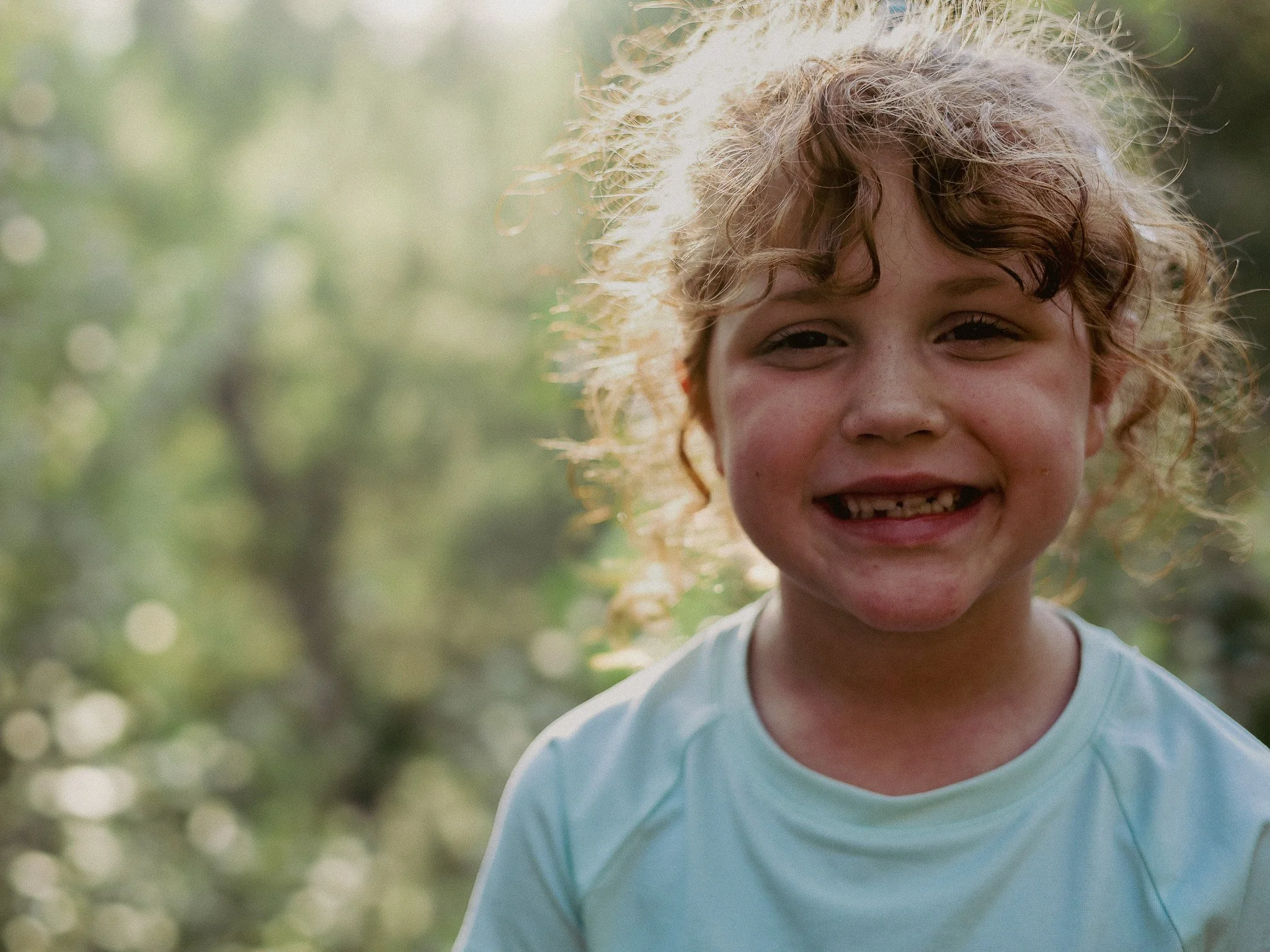 A smiling young girl with curly hair outdoors in natural light, wearing a light-colored shirt, with a blurred green background.