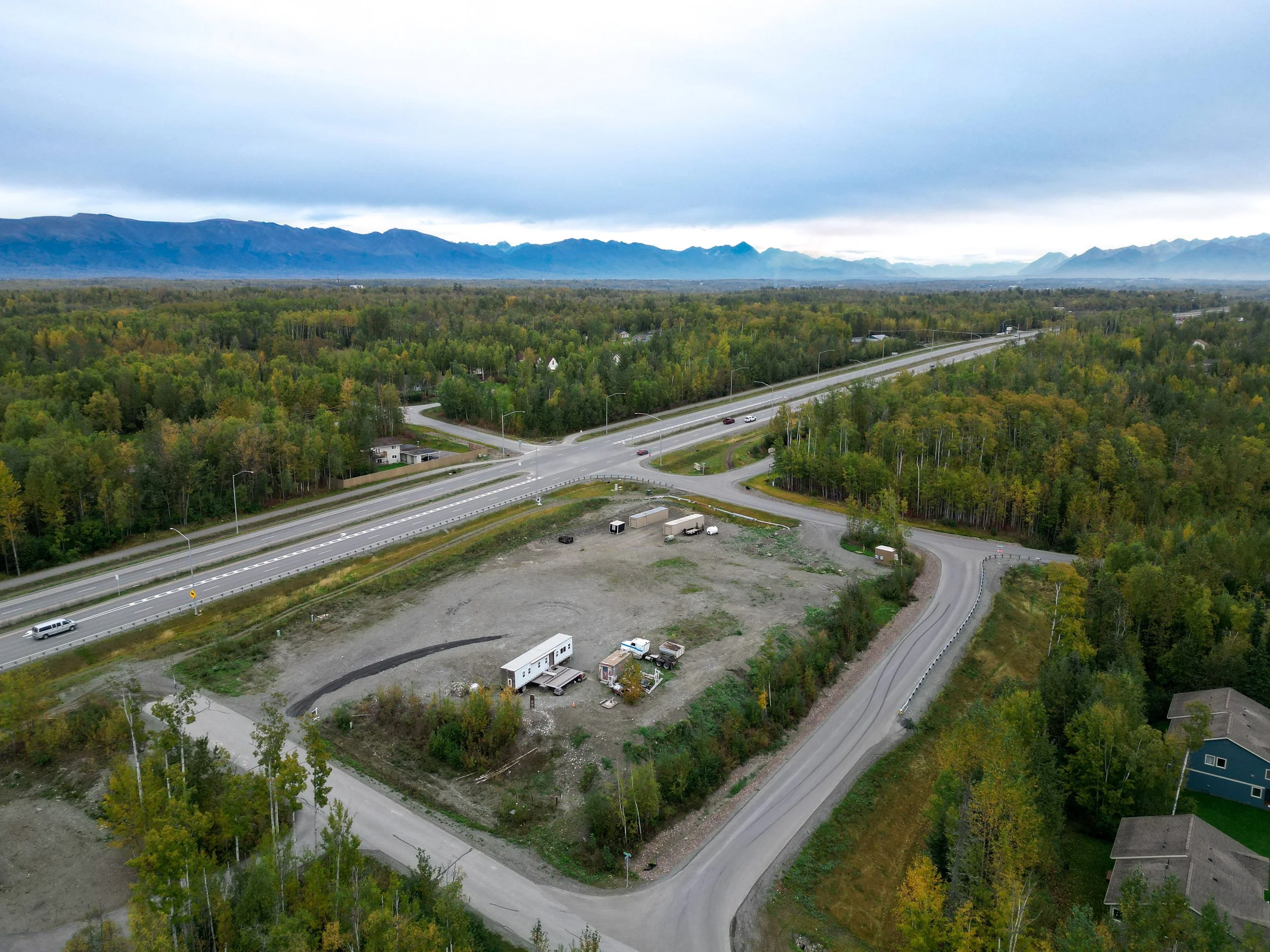 An aerial view of a highway intersection surrounded by trees and residential houses in a mountainous region under cloudy sky.