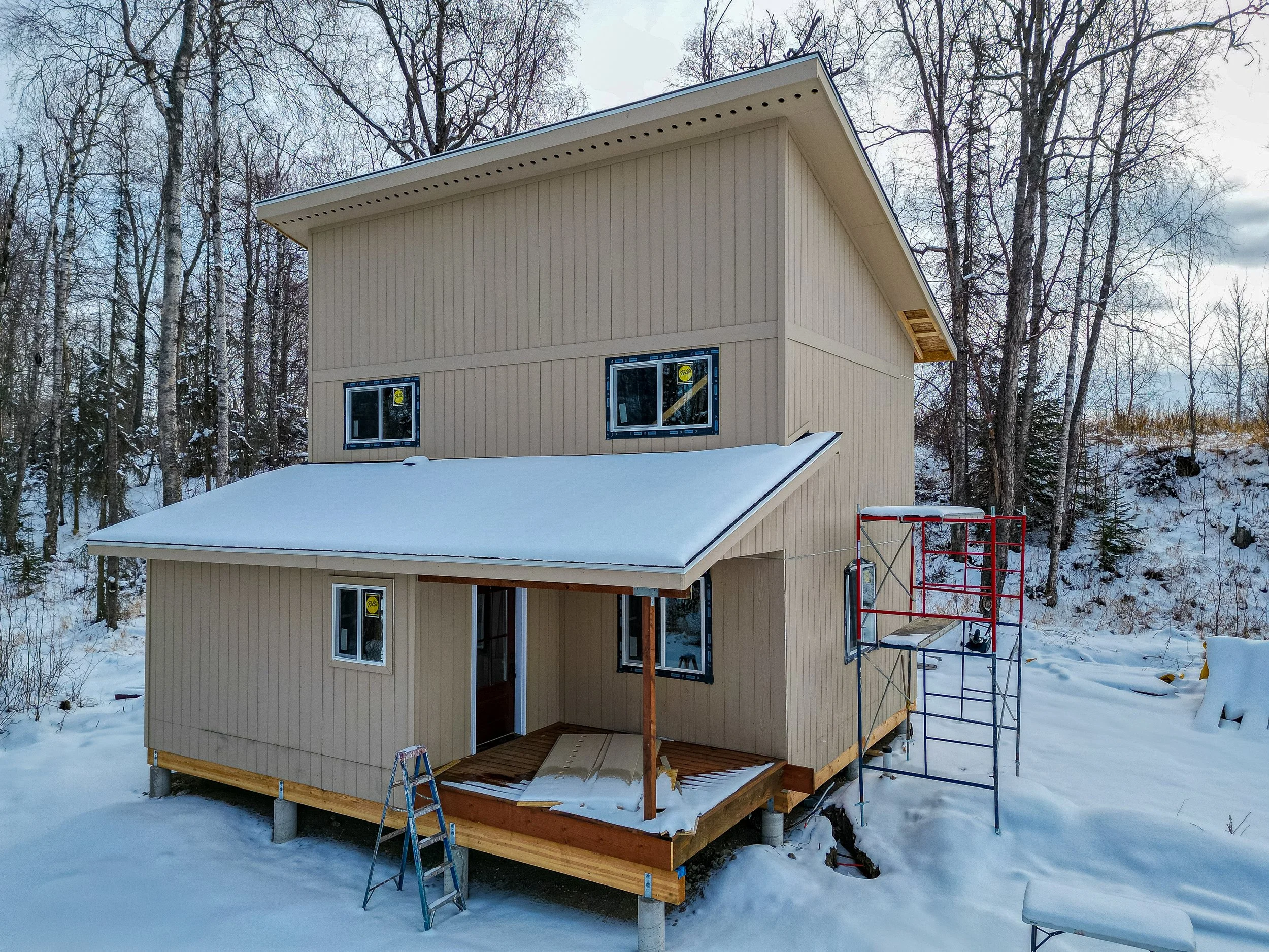 Under construction two-story beige house with scaffolding, located in snowy landscape surrounded by leafless trees, with a wooden deck and partially installed windows.