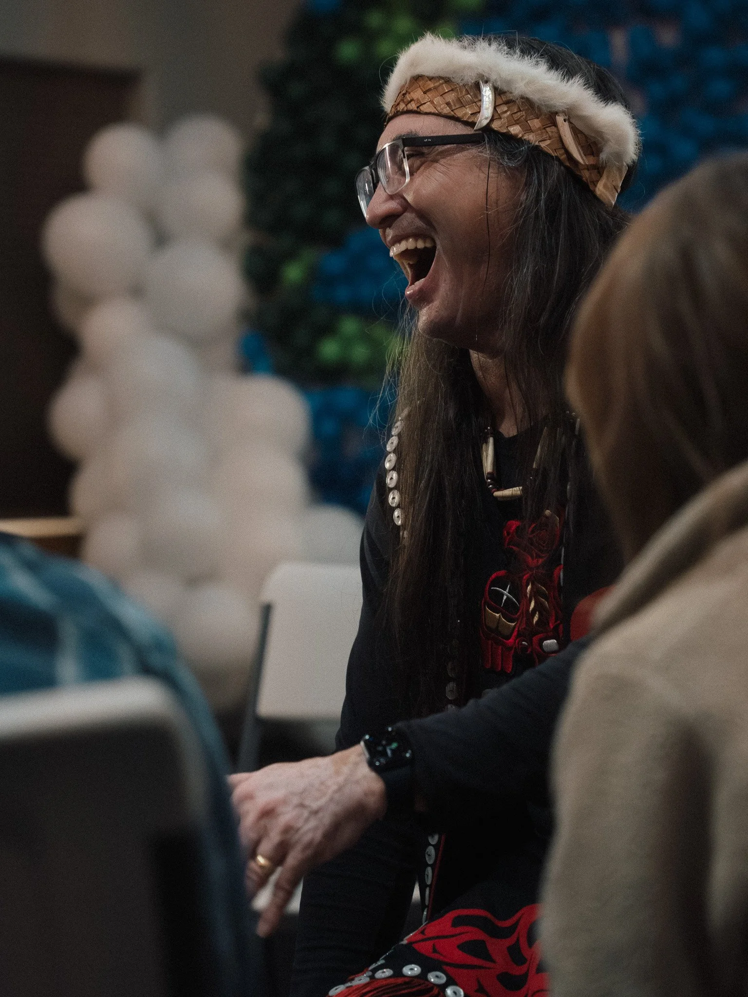 A woman wearing glasses, a beaded headband with fur trim, and a black shirt with Native American designs, is laughing and engaging in conversation at a social event, with a blurred festive background.