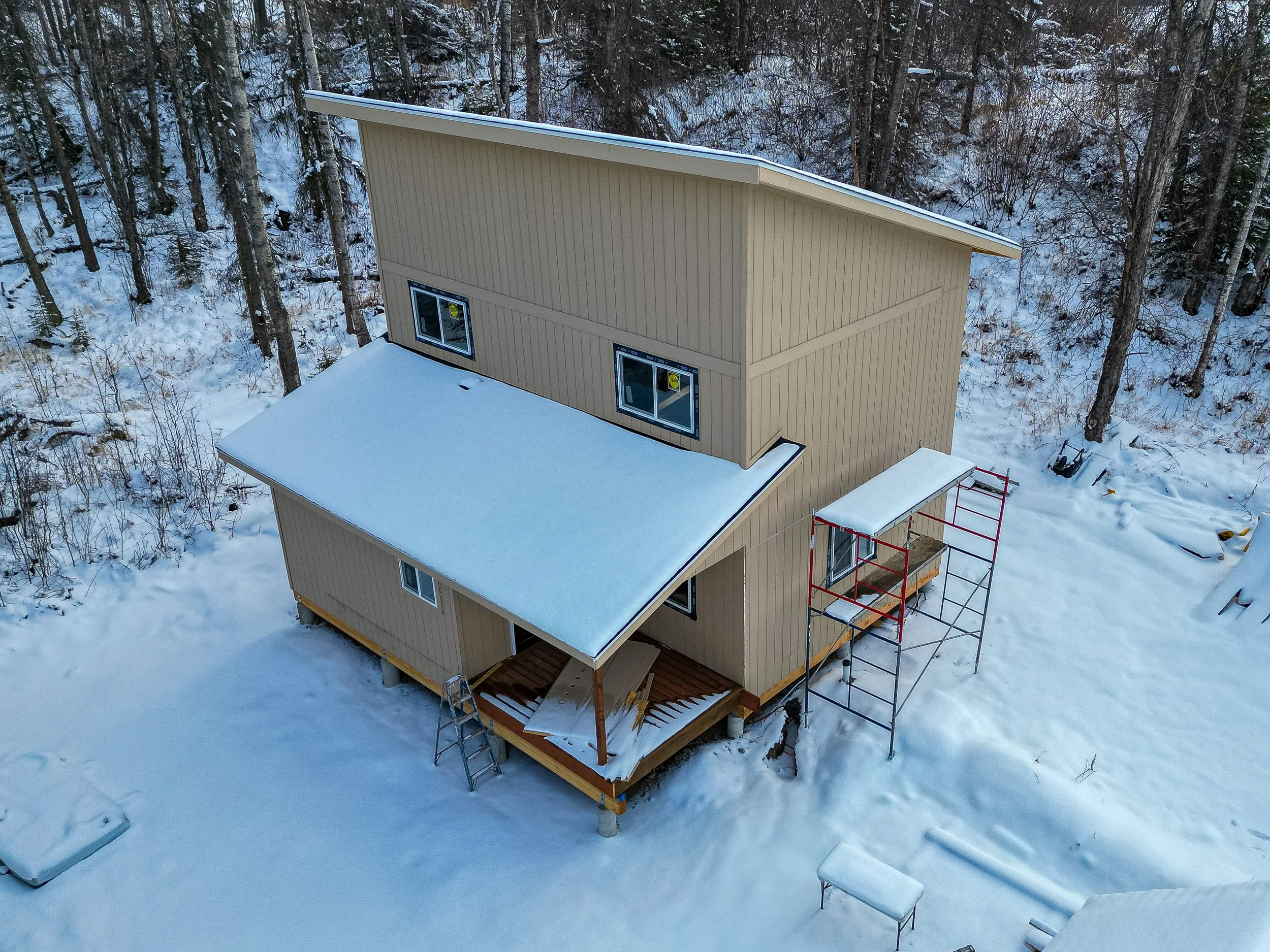 A two-story beige house under construction in a snowy forested area, with scaffolding on one side and snow covering the roof and porch.