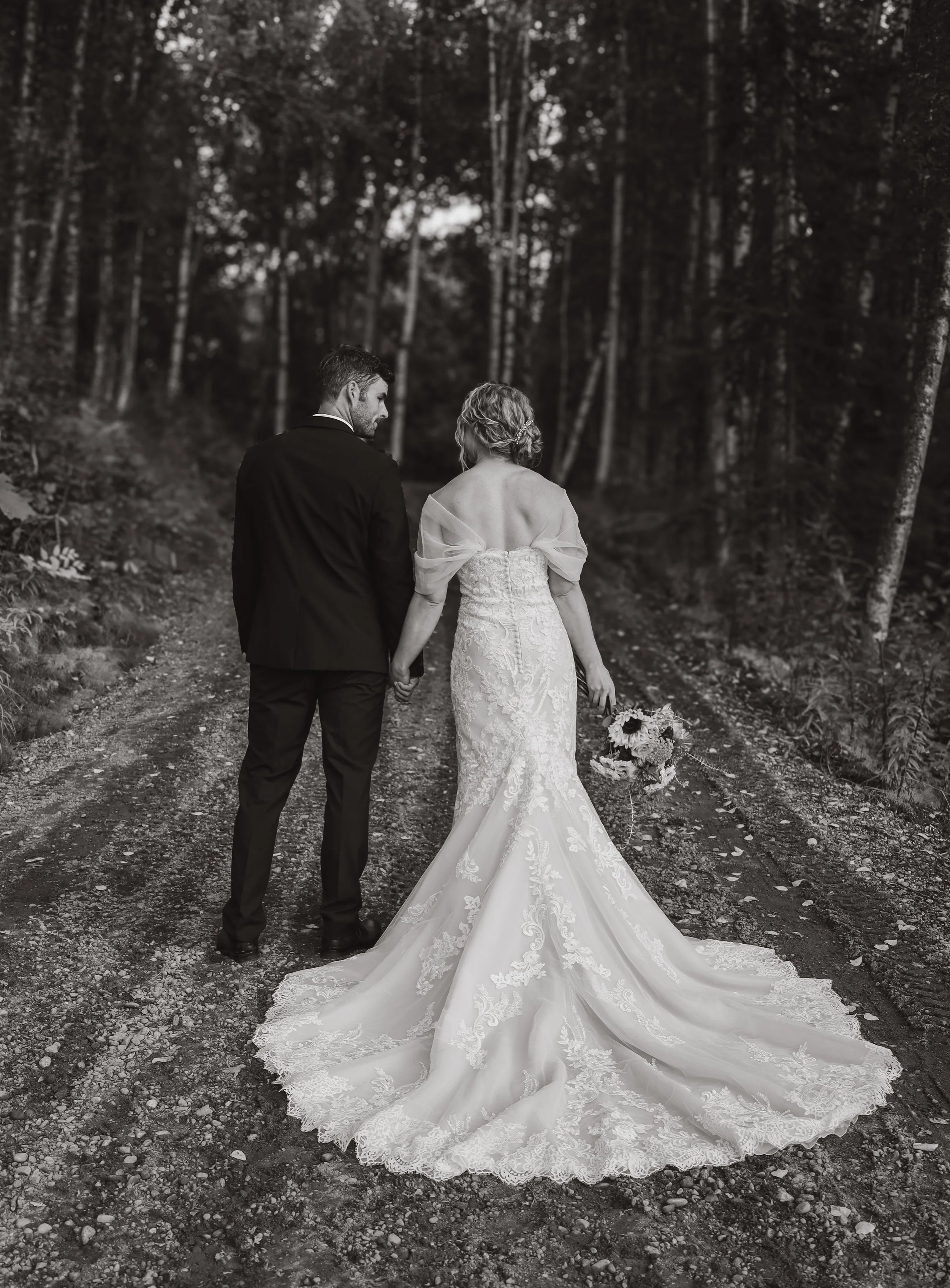 A bride and groom walking hand in hand along a forest trail, with the bride holding a bouquet, as seen from behind in black and white.