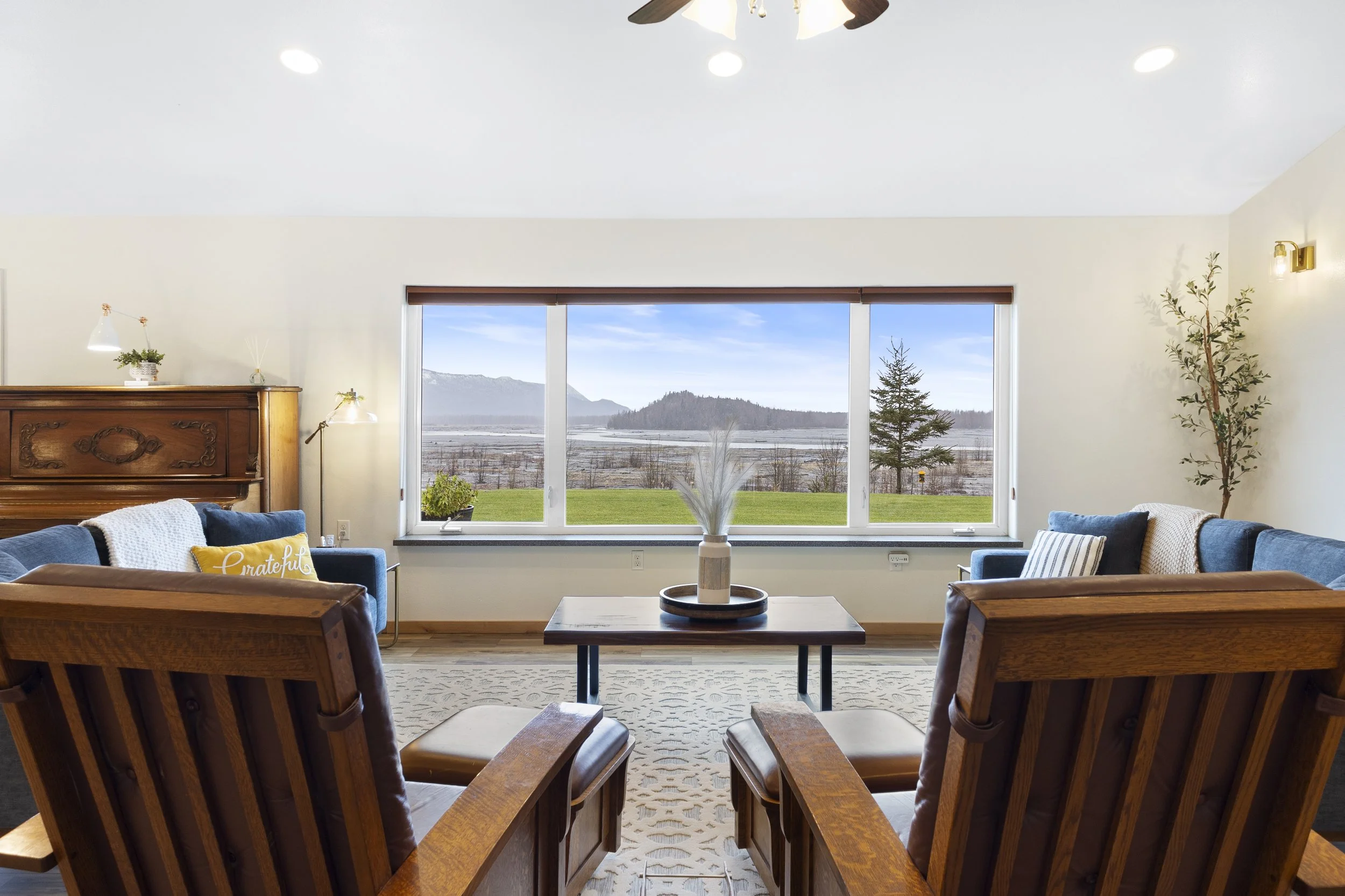 Living room with a large window overlooking a scenic landscape of mountains, trees, and a lake. The room has two blue sofas, a wooden coffee table with a vase, and wooden chairs facing the window. Decor includes a potted plant, wall sconce, and a piano in the corner.