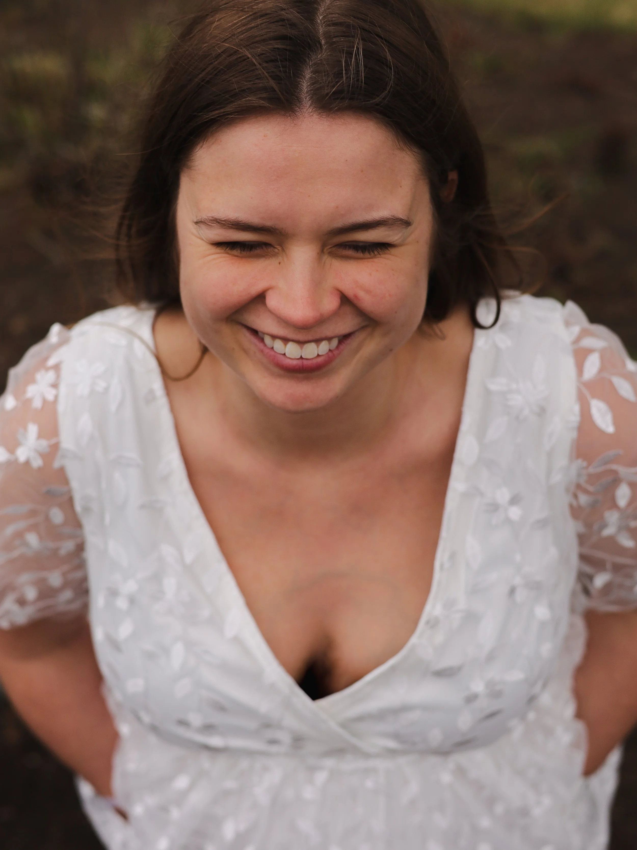 A woman with shoulder-length brown hair smiling with her eyes closed, wearing a white dress with floral embroidery, outdoors on a natural background.