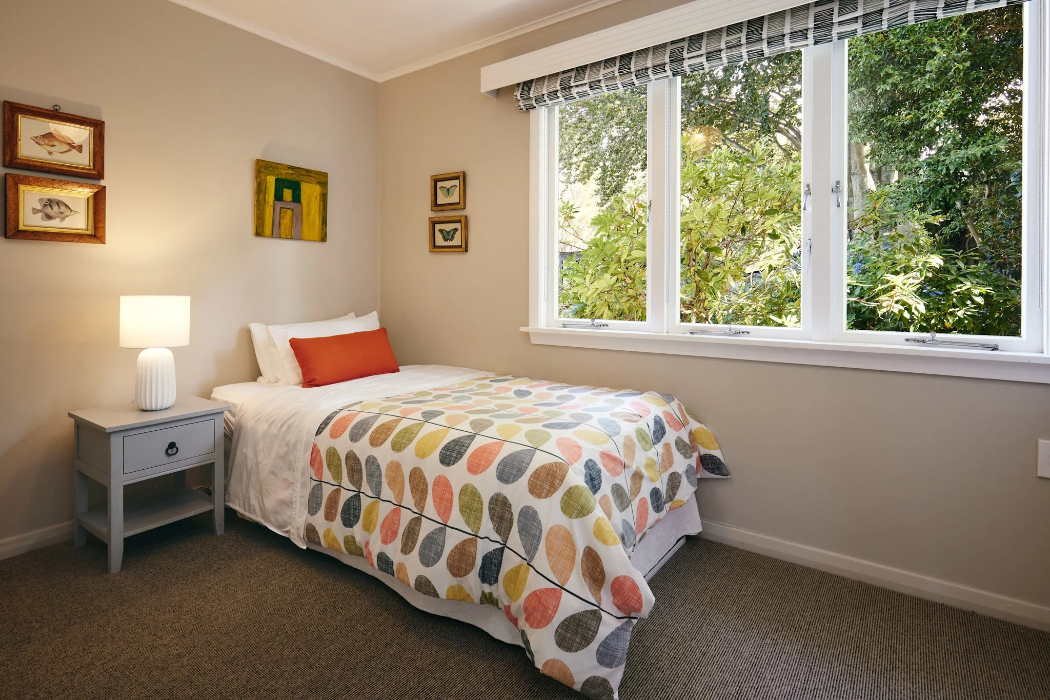 A cozy bedroom with a bed covered in a colorful leaf-patterned quilt, a red pillow, and a white lampshade on a light gray nightstand. There are various framed artworks on the beige walls, and large windows with striped valances revealing green foliag