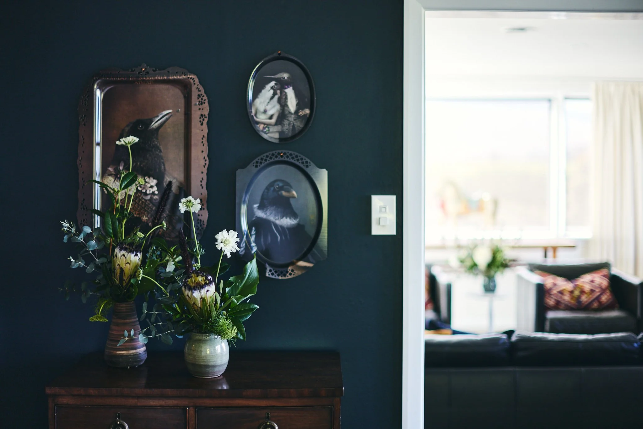 A dark-colored wall decorated with framed bird portraits and a potted flower arrangement on a wooden table, with a brighter room visible through an open doorway in the background.