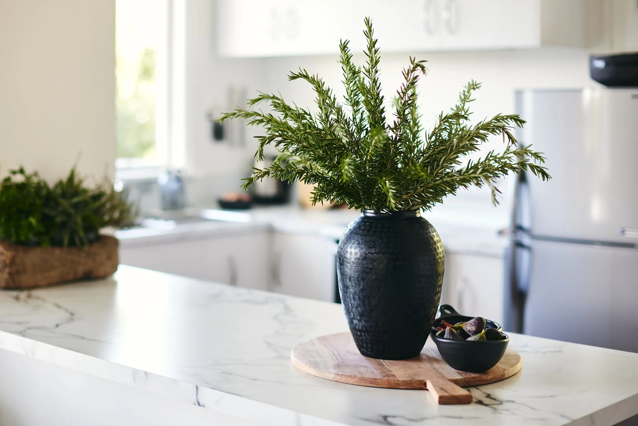 A black vase with green leafy plants on a wooden tray on a white marble kitchen countertop.