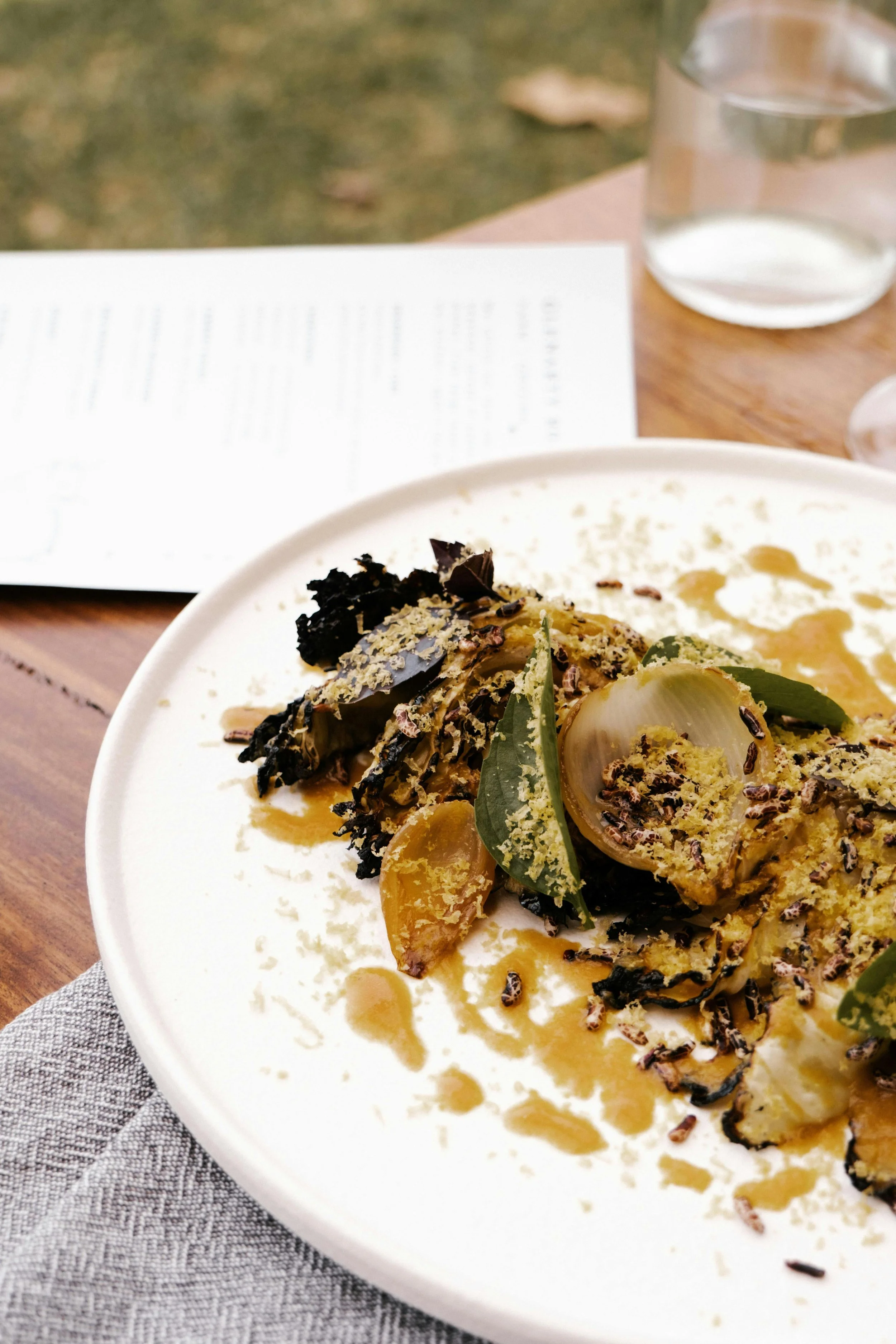 Close-up of a plated gourmet dish with grilled vegetables, herbs, and sauce on a white plate, set on a wooden table with a glass of water and a menu in the background.