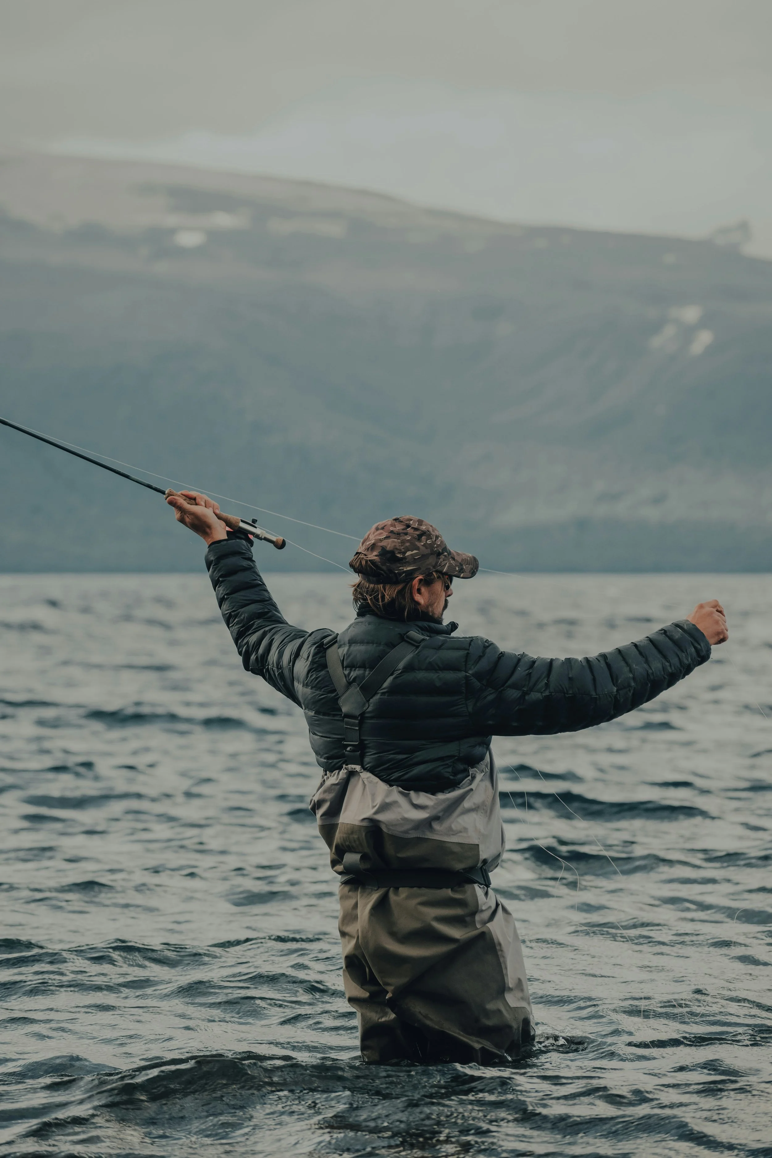 Person fishing in a lake with mountains in the background, wearing a black jacket, brown waders, and a camouflage hat.