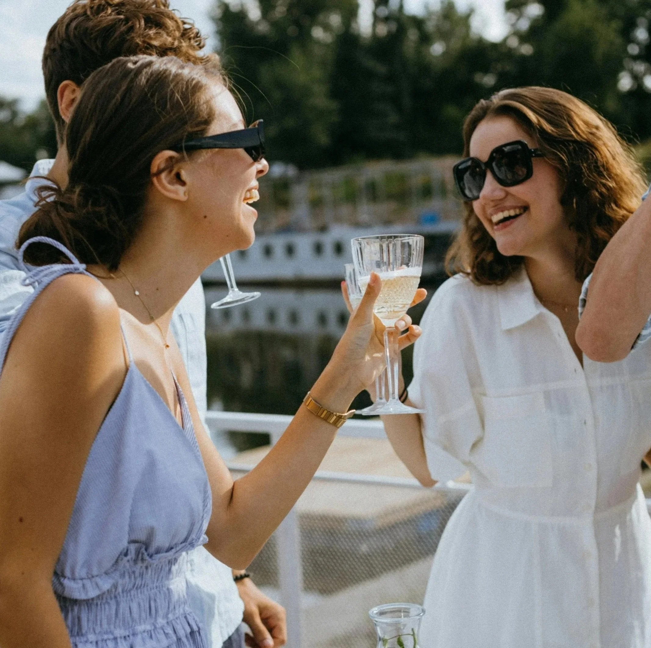 A group of women enjoying a drink and laughing together outdoors near a body of water.