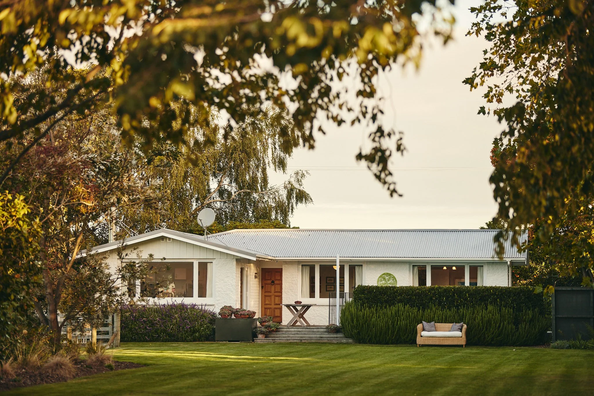 A white house with a metal roof, surrounded by greenery and trees, with a small outdoor seating area on the front porch.