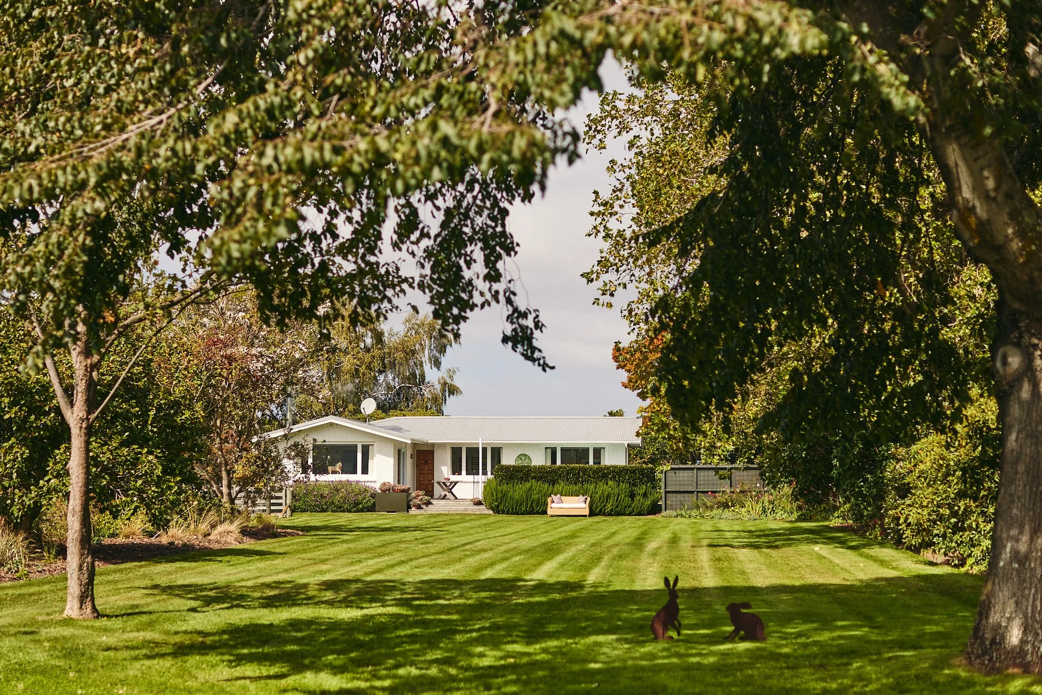 A white house with a gray roof in the background, surrounded by green trees and bushes, with a well-maintained lawn and two bunny garden statues in the foreground.