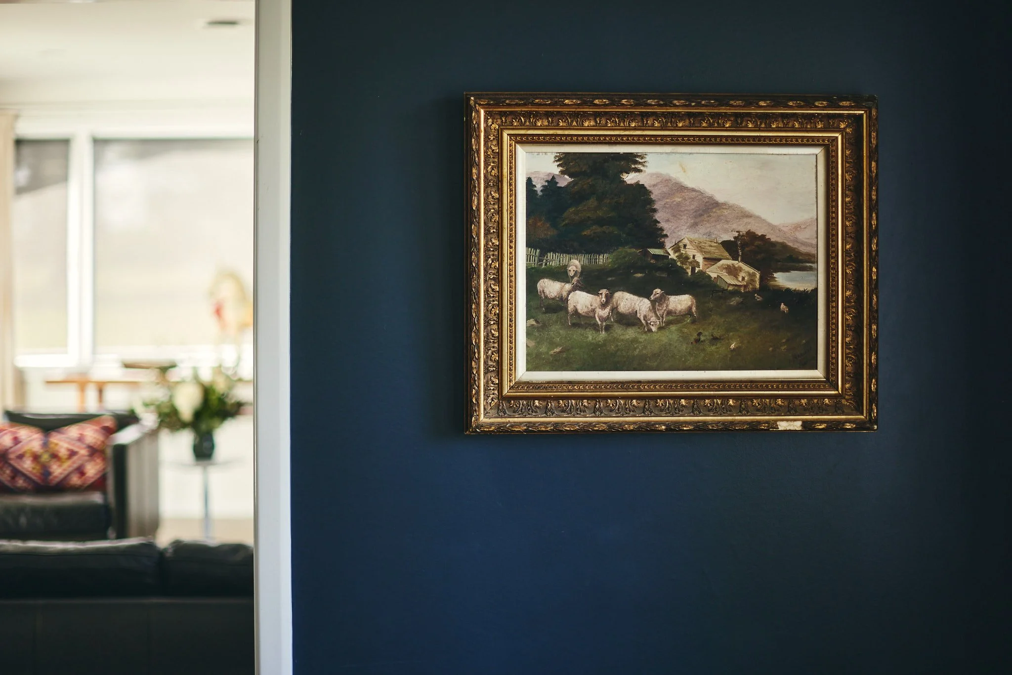 A framed landscape painting of sheep grazing near a rural house, with trees and mountains in the background, hanging on a dark blue wall.