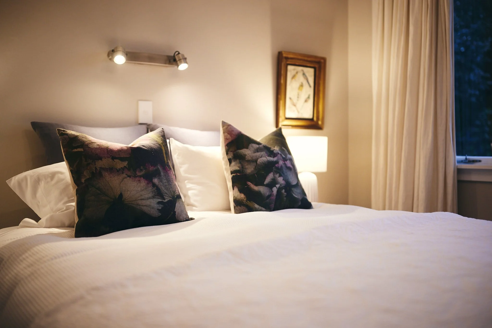 A neatly made bed with white bedding and three decorative pillows with purple and black floral patterns in a cozy bedroom with beige walls, a wall-mounted reading light, a framed art piece, and a window with cream-colored curtains.