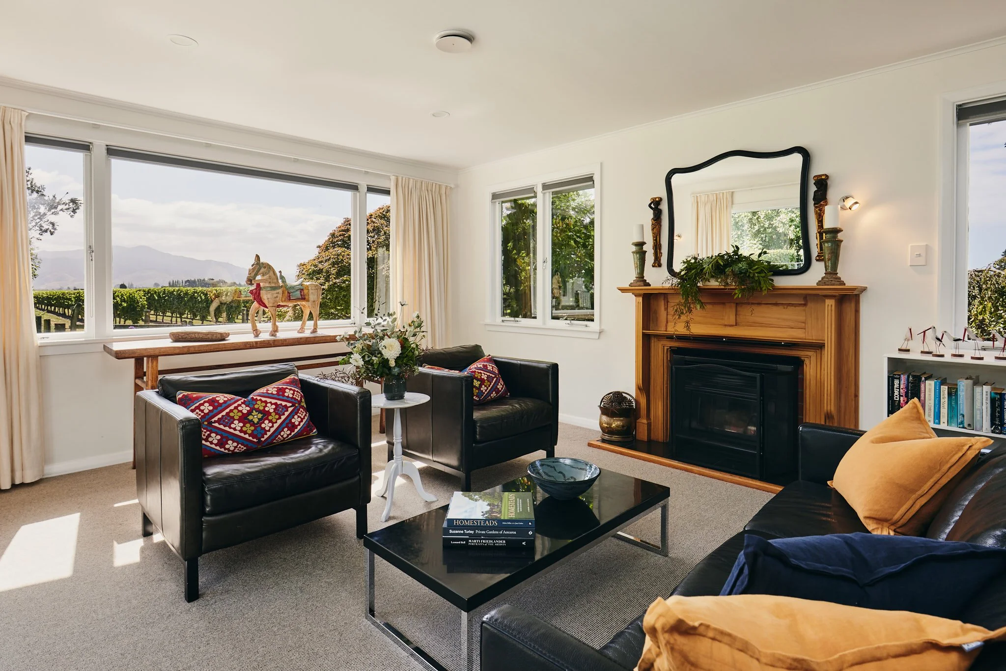 Living room with black sofas, a wooden fireplace, a large window with a view of trees and mountains, and decorative items including a rocking horse and books.