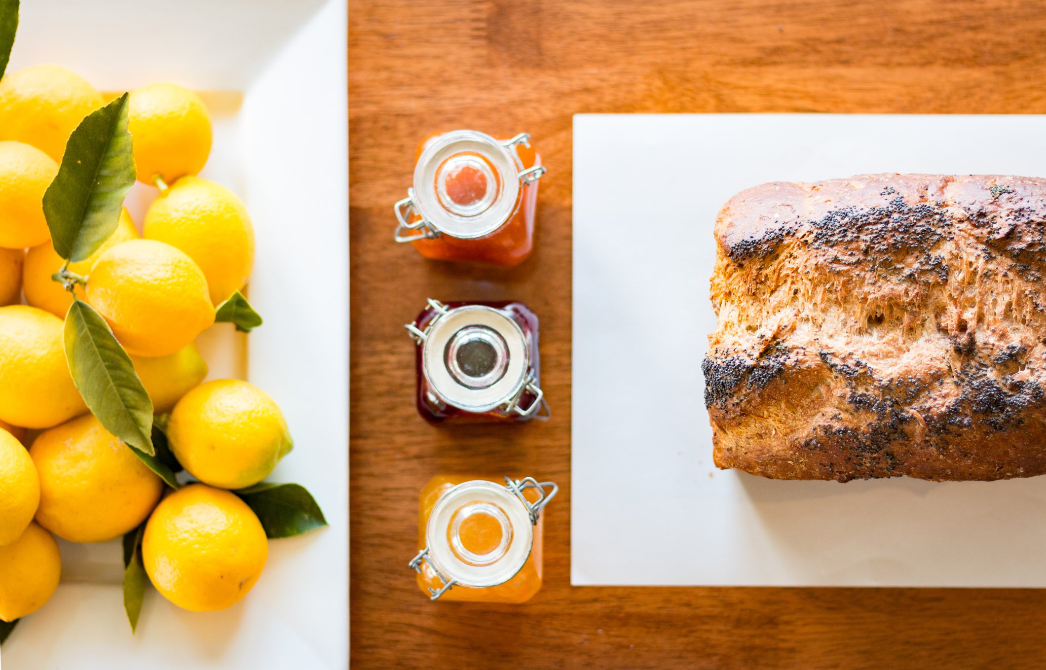 A loaf of bread on a white cutting board, with three small jars of jam or preserves nearby, and a bunch of yellow lemons with green leaves on a white plate.