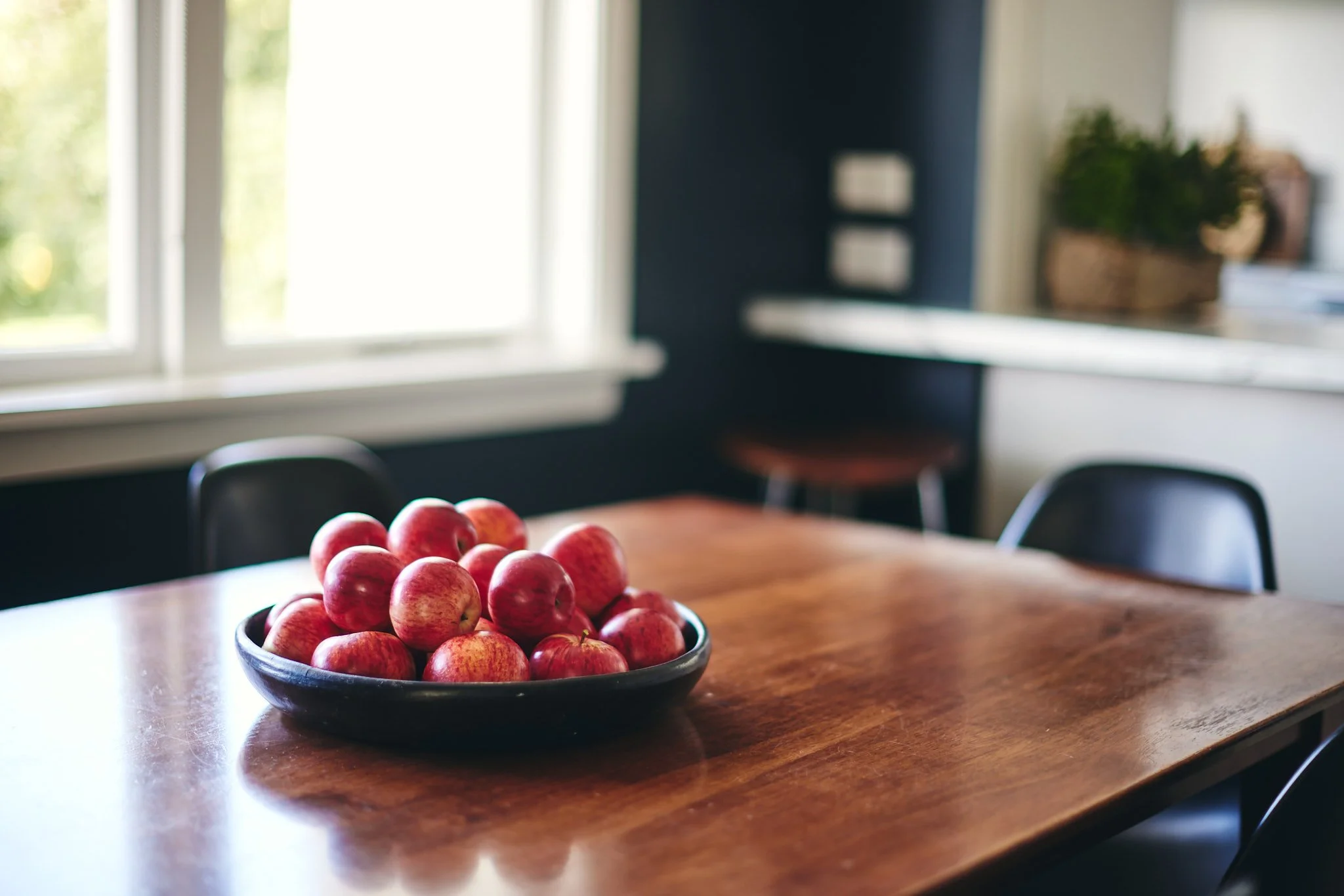 A black bowl filled with red apples on a wooden dining table in a brightly lit kitchen.