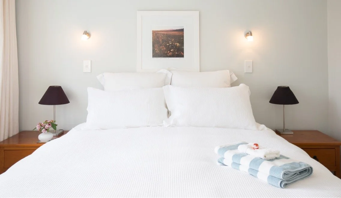 Clean bedroom with white bedding, two nightstands with black lamps, a framed landscape photo above the bed, and folded towels on the bed.